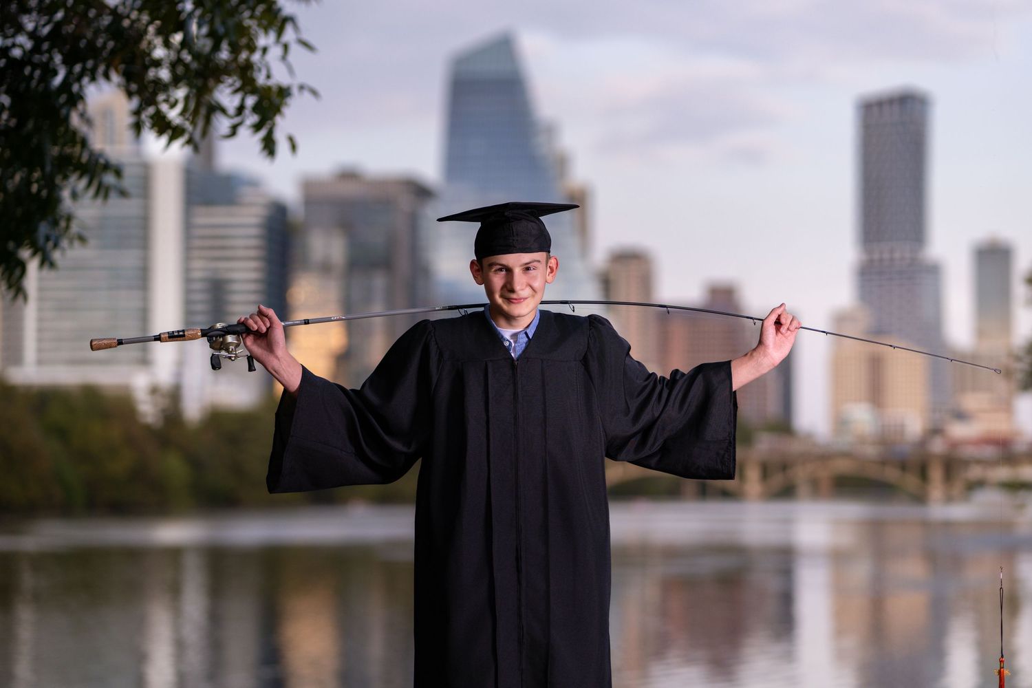 Graduate in cap and gown holds fishing rod outdoors with city skyline in background.