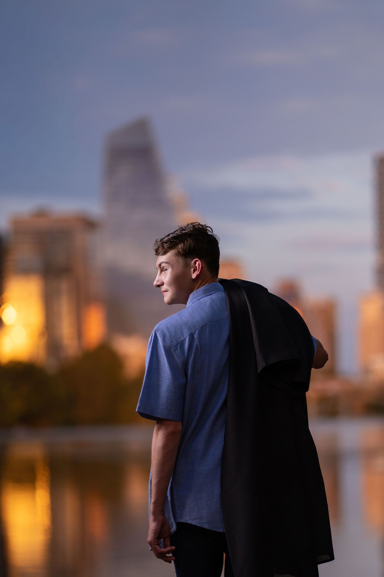 Young man in blue shirt holding jacket stands by urban waterfront at dusk with city skyline reflected in water.
