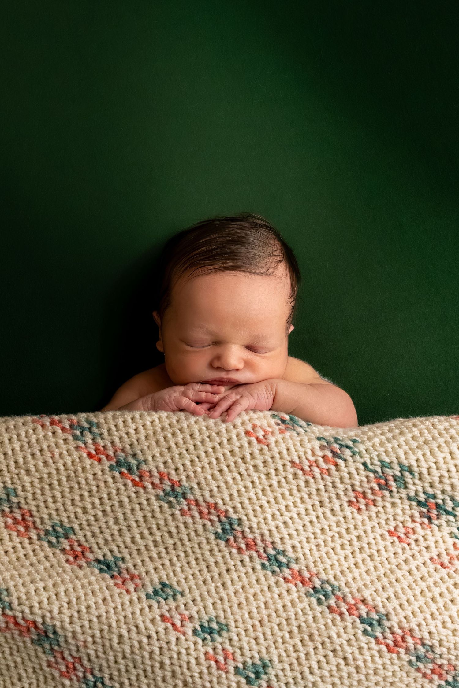Sleeping newborn baby resting chin on hands over a cream knit blanket against a dark green background.