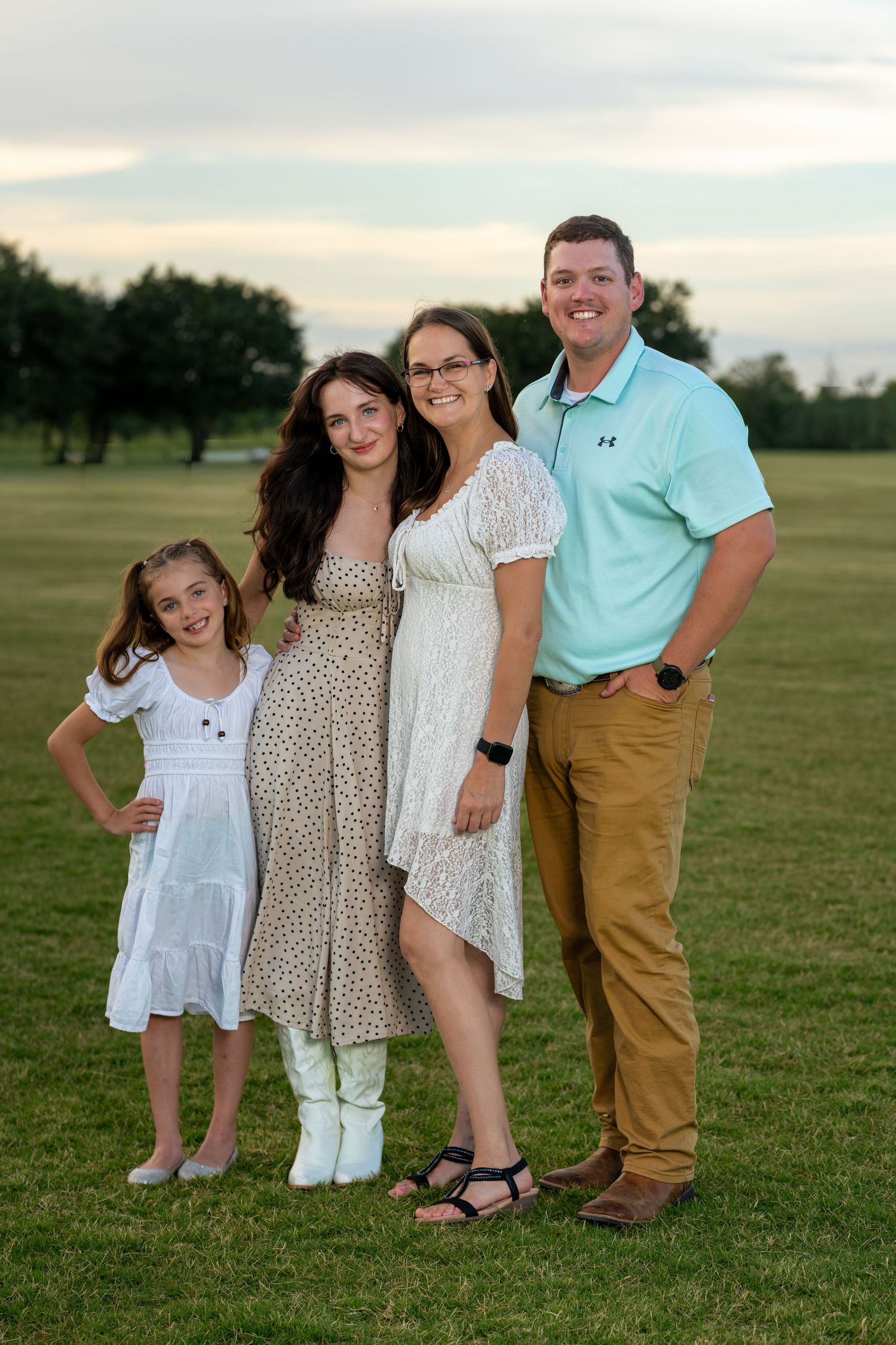 Family of four posing outdoors on green grass at sunset, wearing casual summer attire, smiling together.