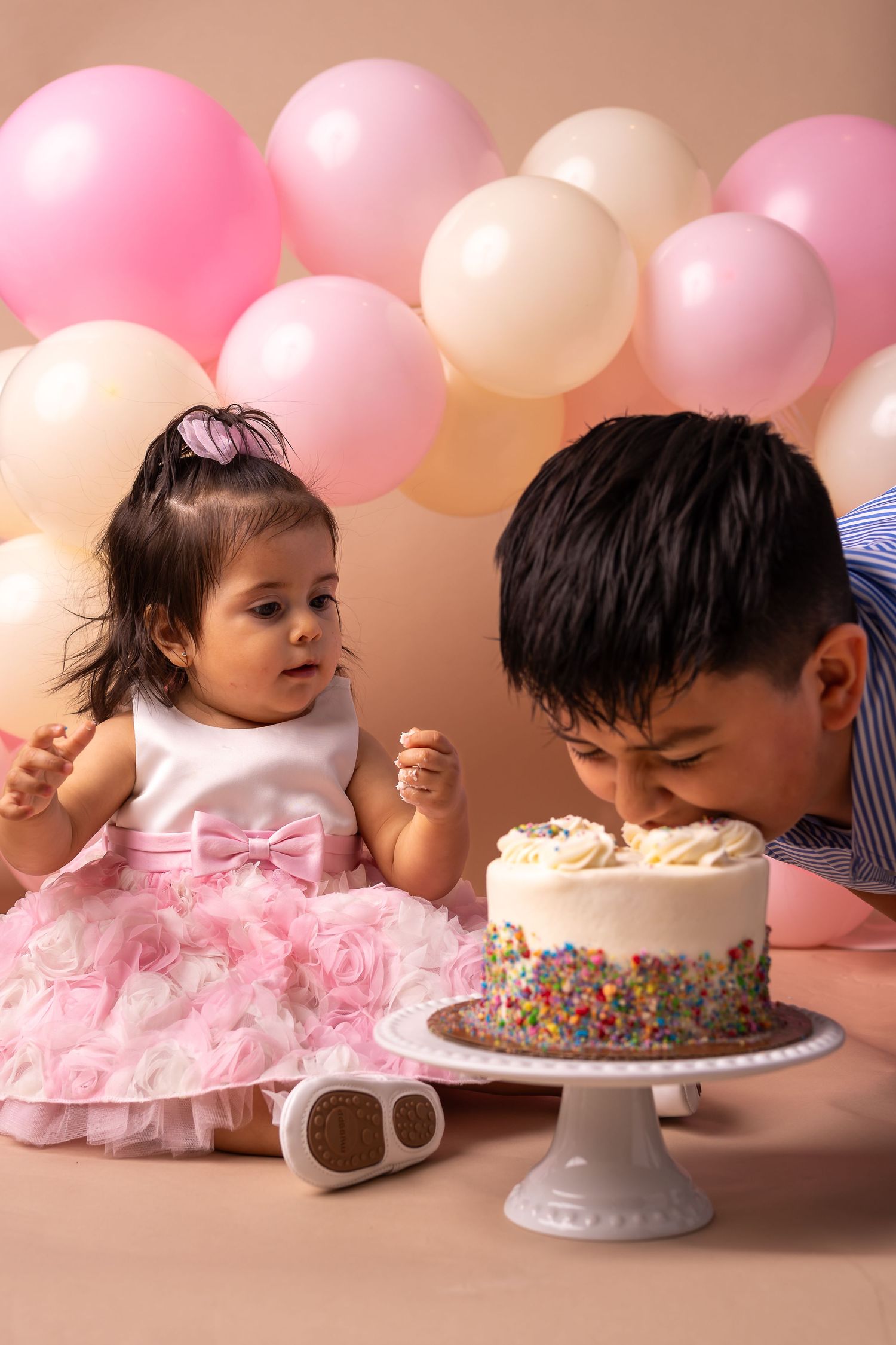 Cake smash photo sequence showing baby in pink dress diving face first into birthday cake with pink balloons in background.