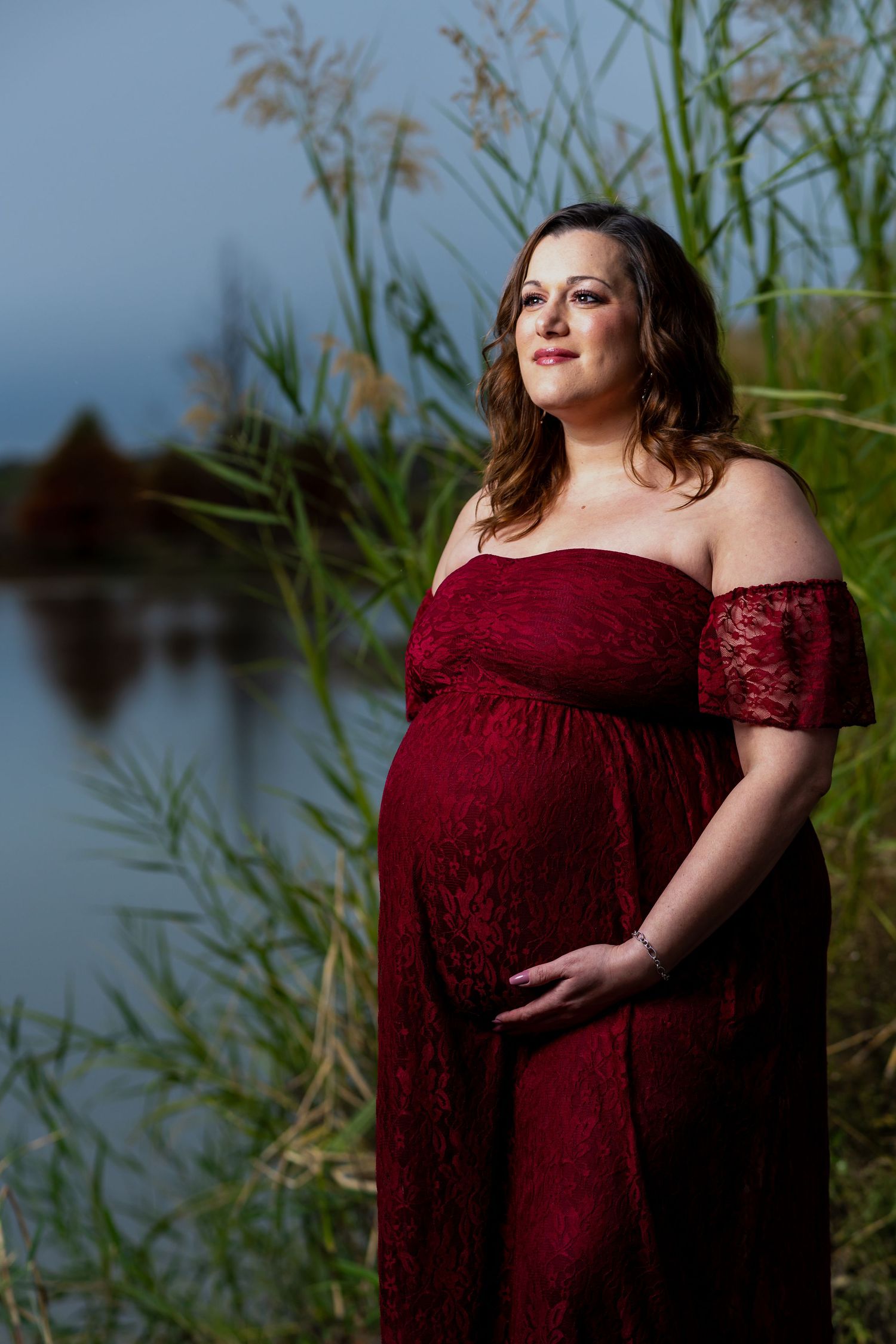 A pregnant woman in a burgundy lace dress poses gracefully by a lake with reeds in the background.