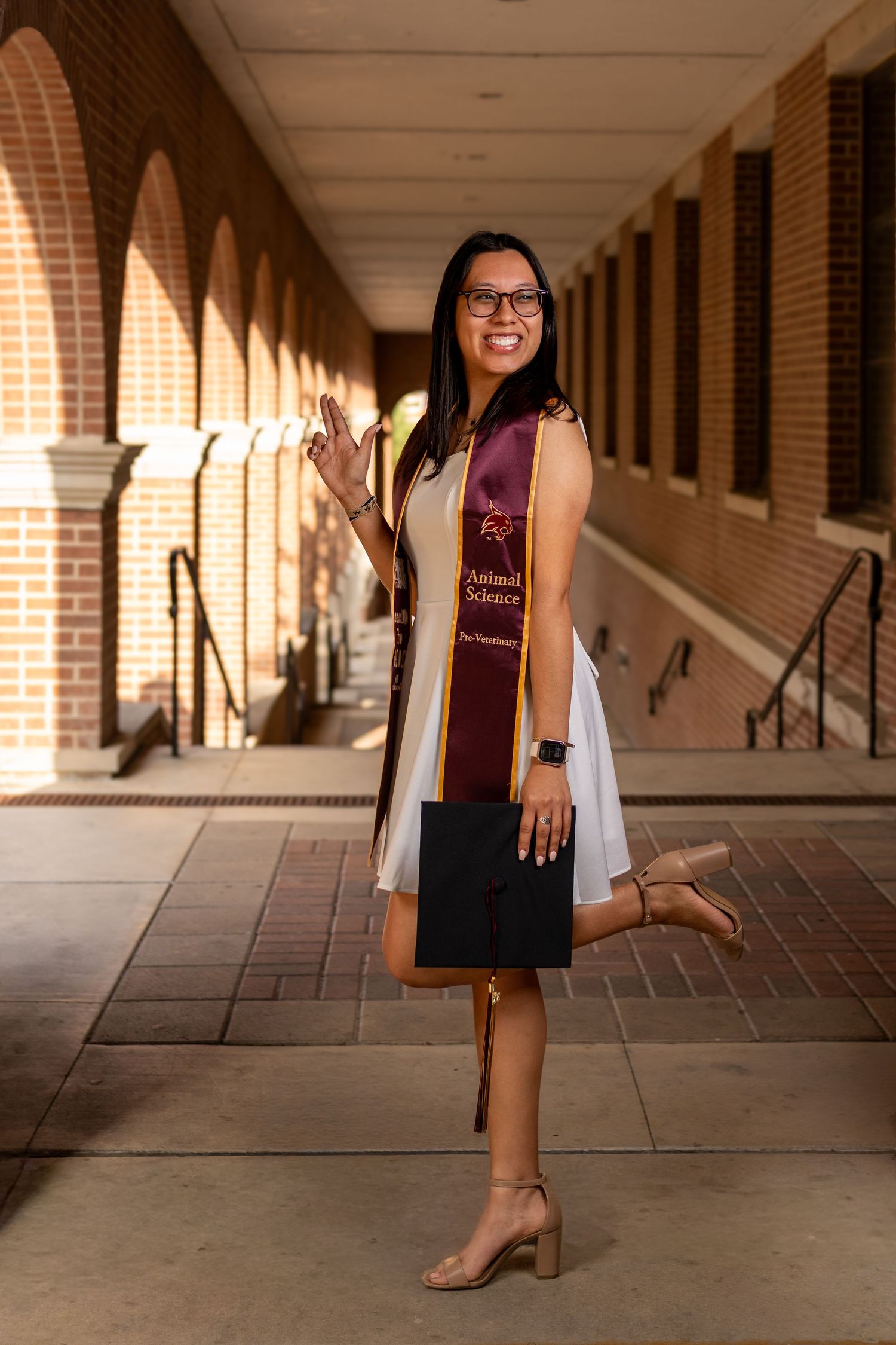 Smiling graduate poses in white dress with maroon ASU stole holding cap in brick archway corridor.