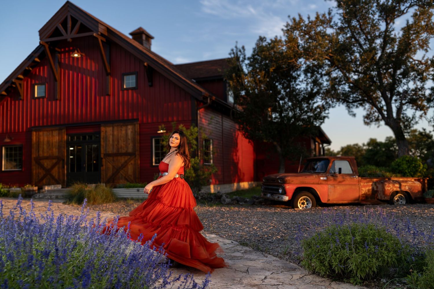 Woman in red gown poses near red barn and vintage orange truck surrounded by lavender at golden hour.