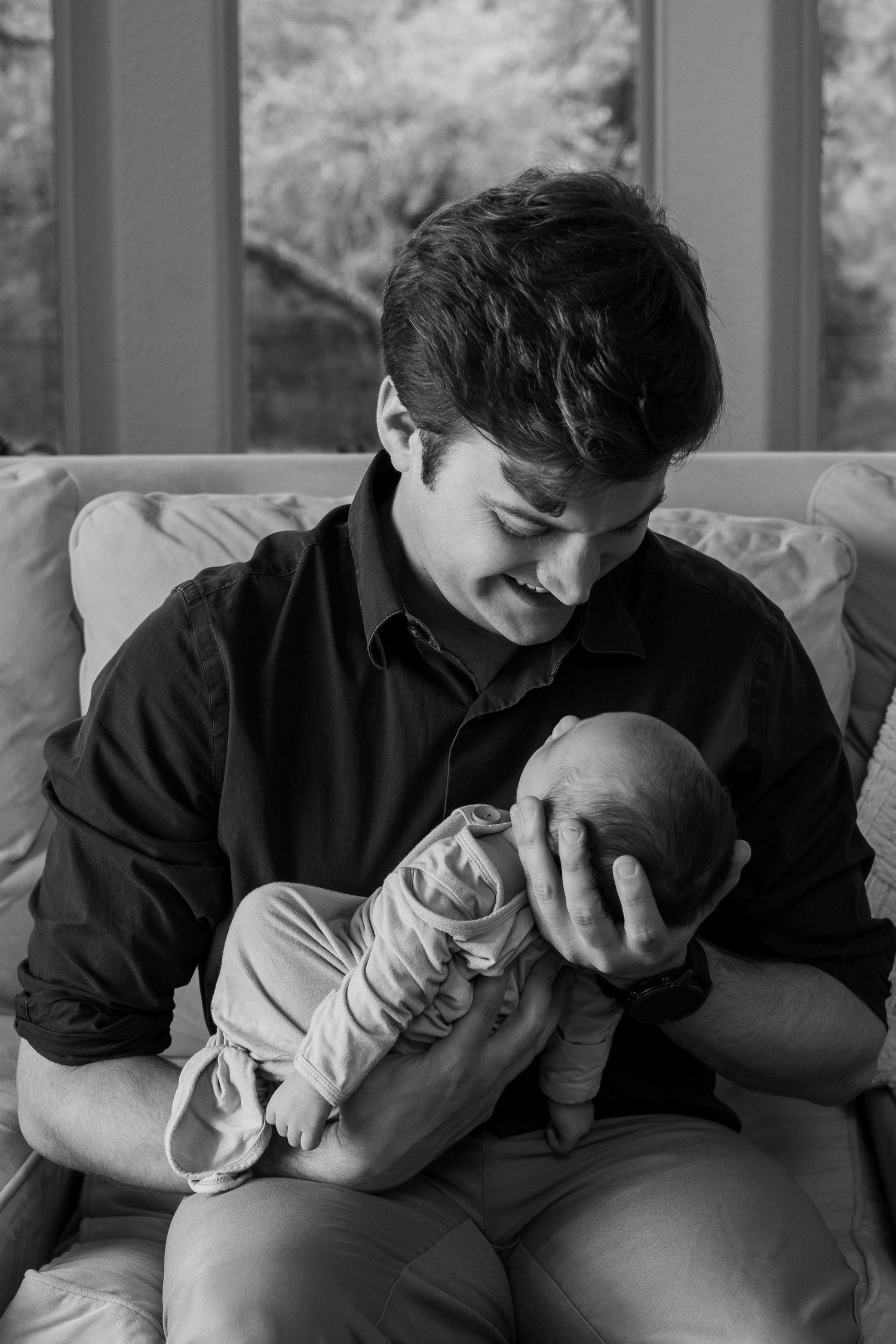 Black and white photo of a young man cradling a newborn baby while sitting on a couch near a window.