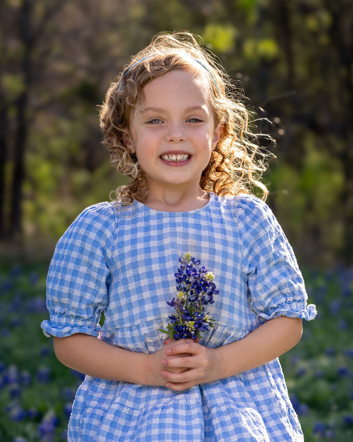 A child in a blue gingham dress holds a small bouquet of wildflowers while standing in a sunlit meadow.