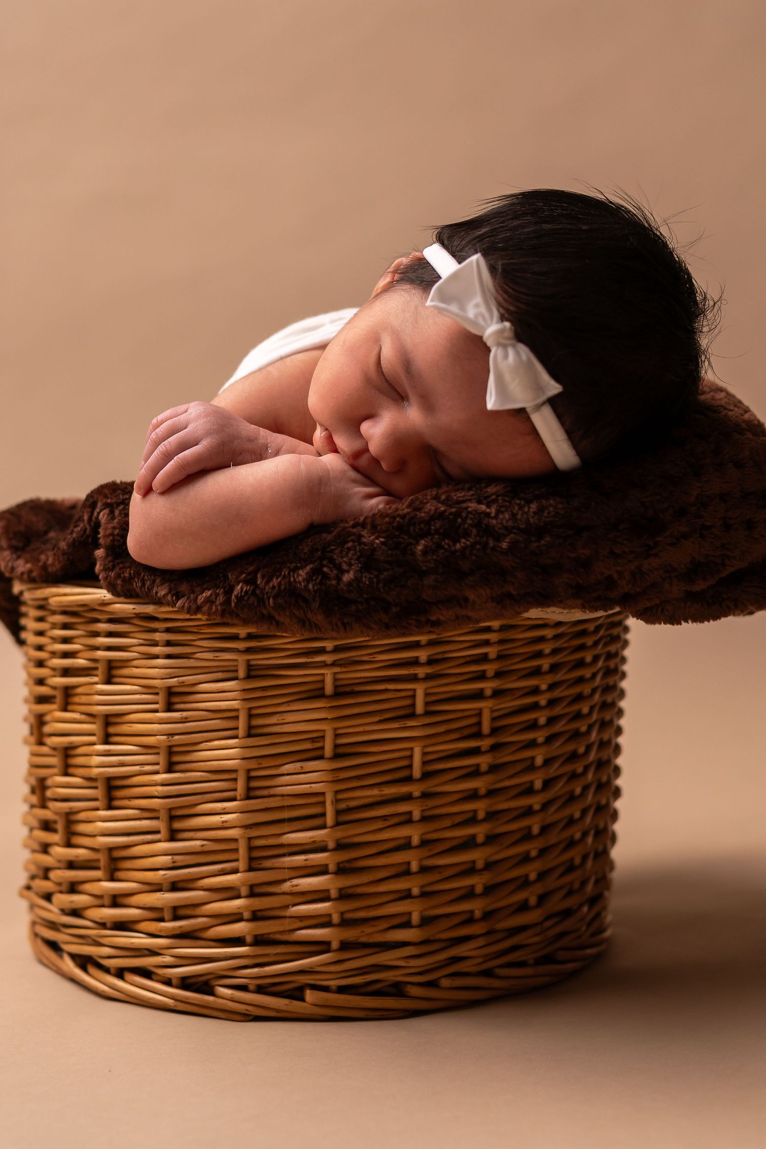 Sleeping newborn rests in woven wicker basket with soft brown backdrop.