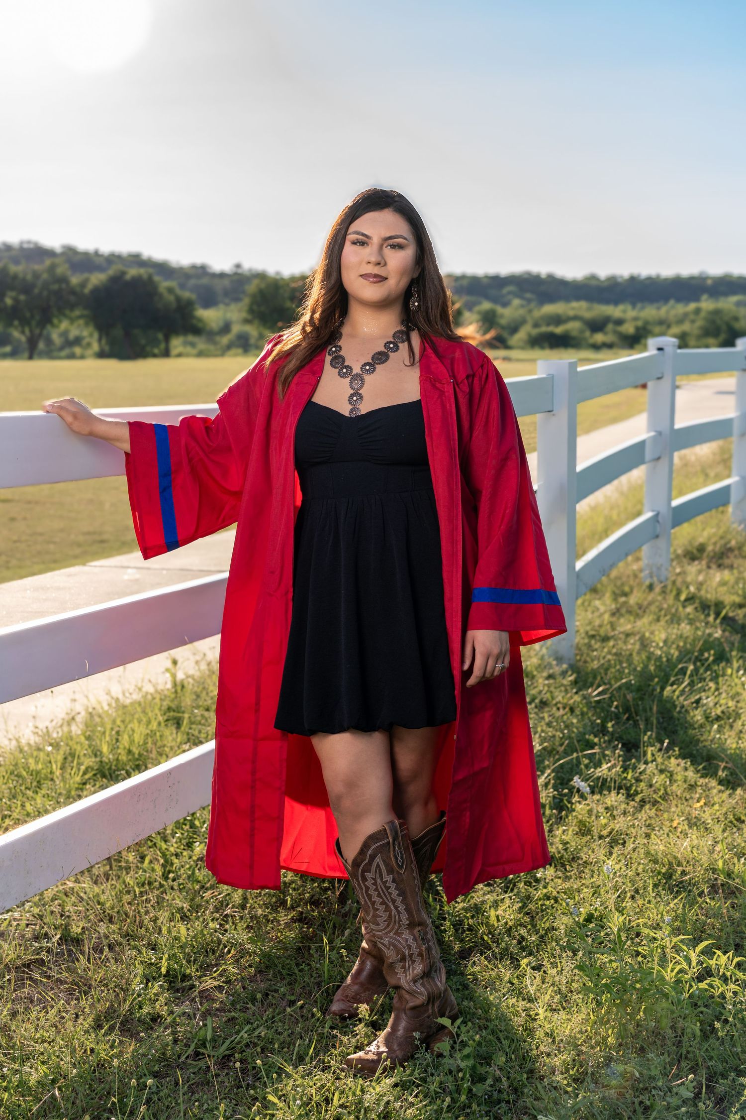 Young woman in red graduation gown and cowboy boots poses by white fence in scenic outdoor setting.