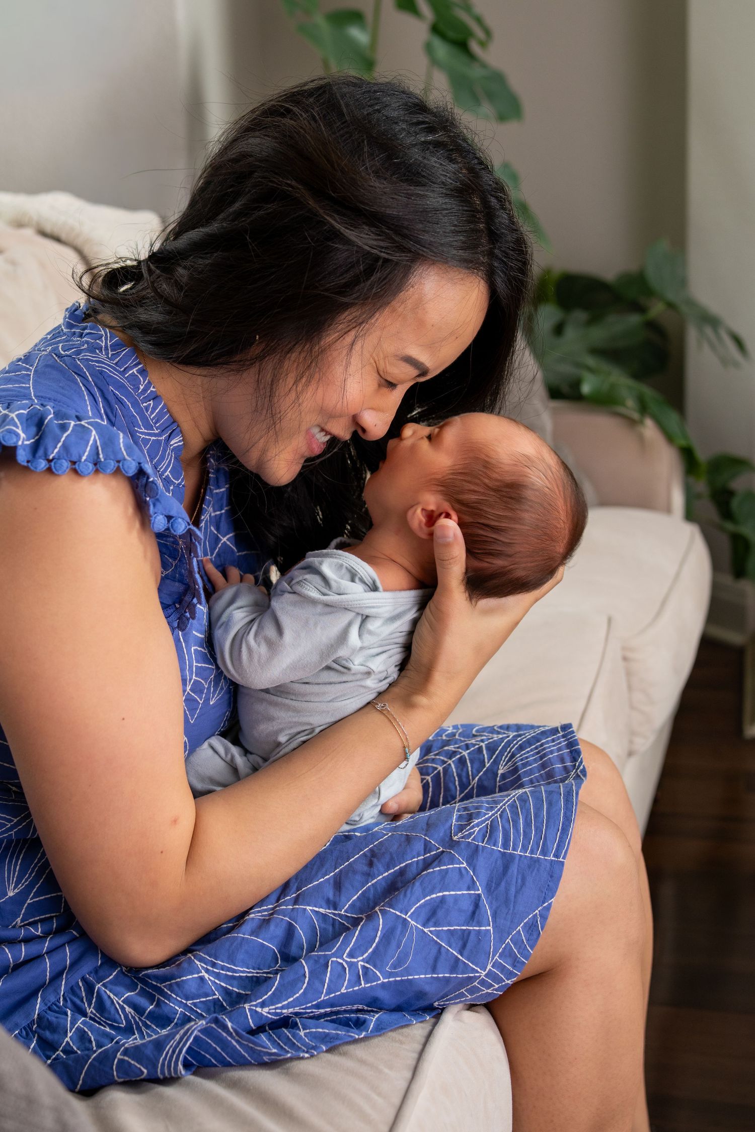 A mother in a blue dress tenderly holds her newborn baby, touching foreheads while seated on a couch.