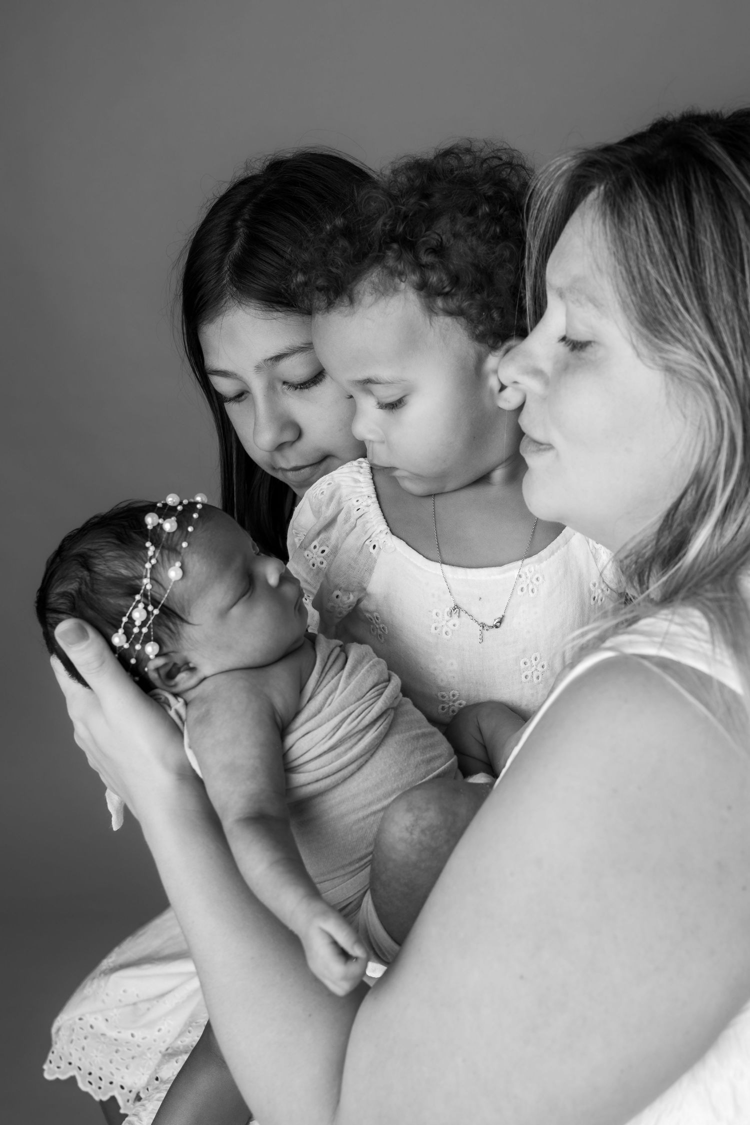 Black and white photo of two women and a young girl lovingly gazing at a sleeping newborn baby with a floral headband.