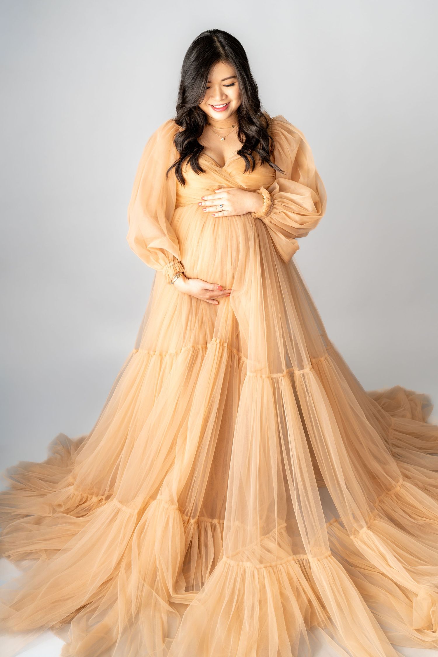 Pregnant woman in elegant gold tulle maternity gown cradling her baby bump in a studio photo.