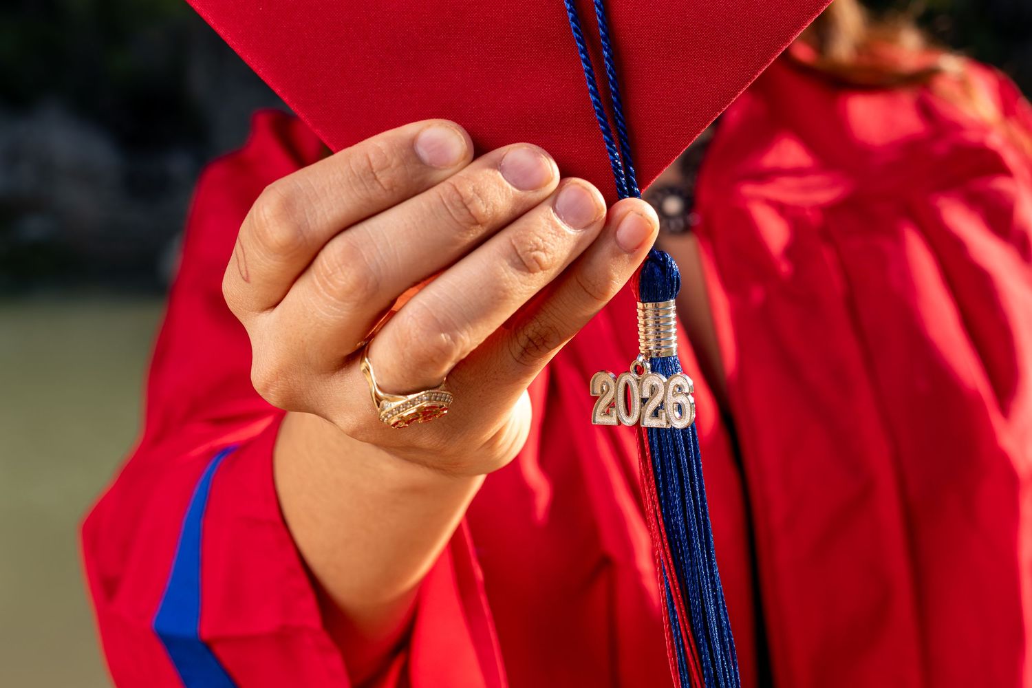 Close-up of a hand holding a red graduation cap with a 2026 tassel charm, wearing a red graduation gown.