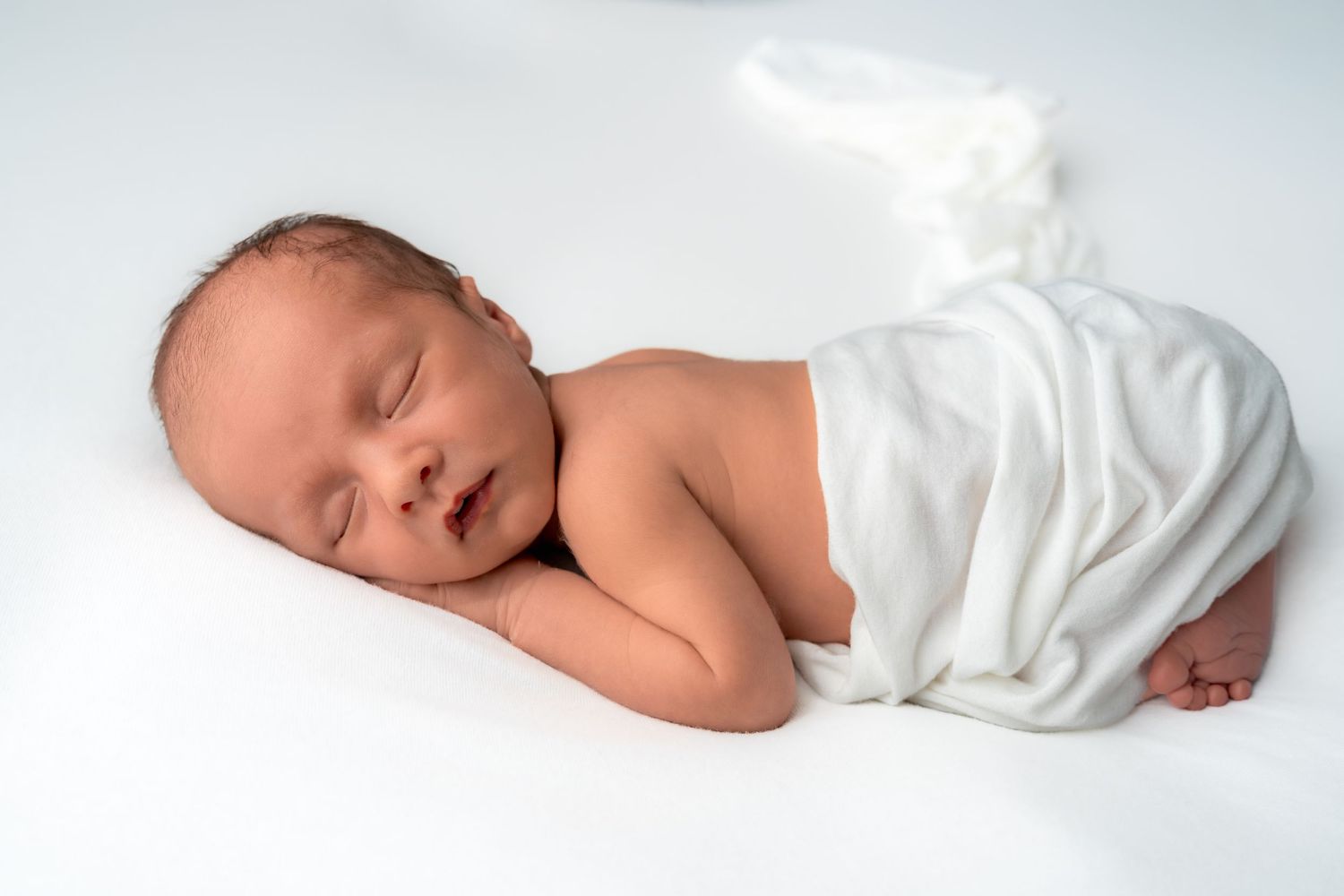 Peaceful newborn baby sleeping wrapped in white cloth on white background.
