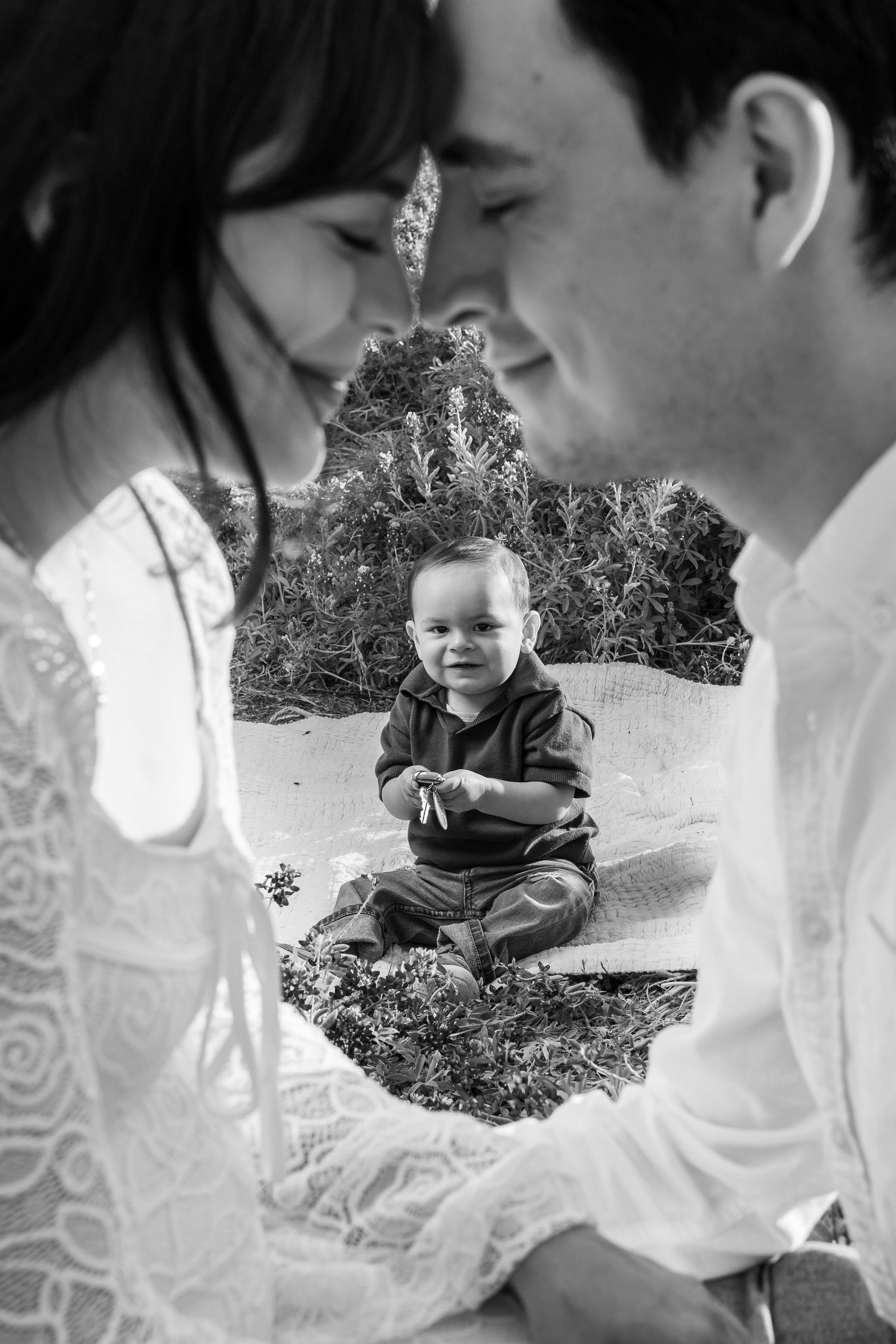Black and white photo of parents leaning in close while baby sits between them outdoors.