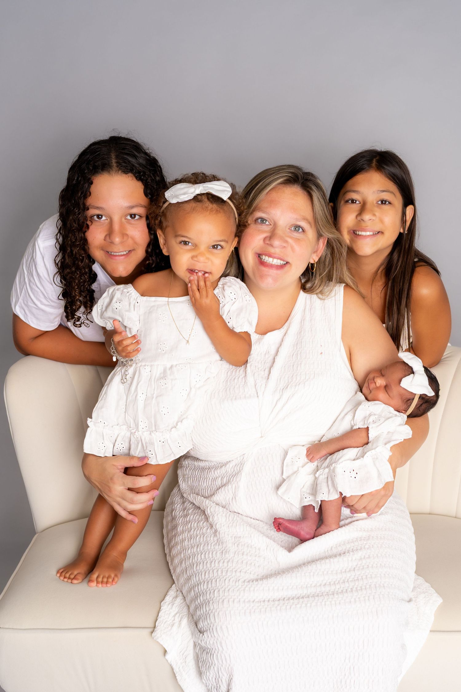 A smiling mother in white holds a newborn while seated with three daughters in a bright studio portrait.