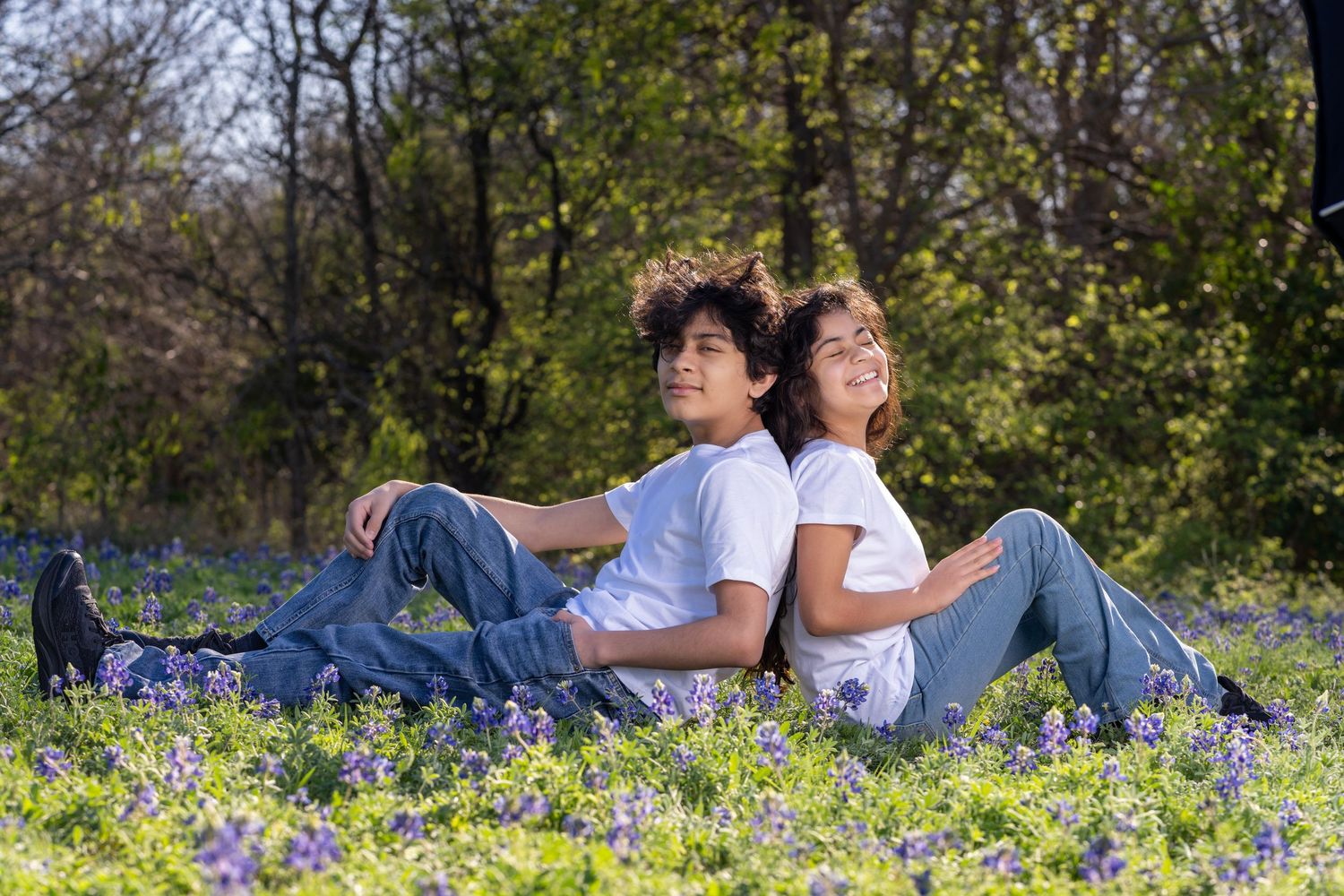 Two people sitting back-to-back in casual wear among blue wildflowers in a sunny outdoor setting with trees in background.