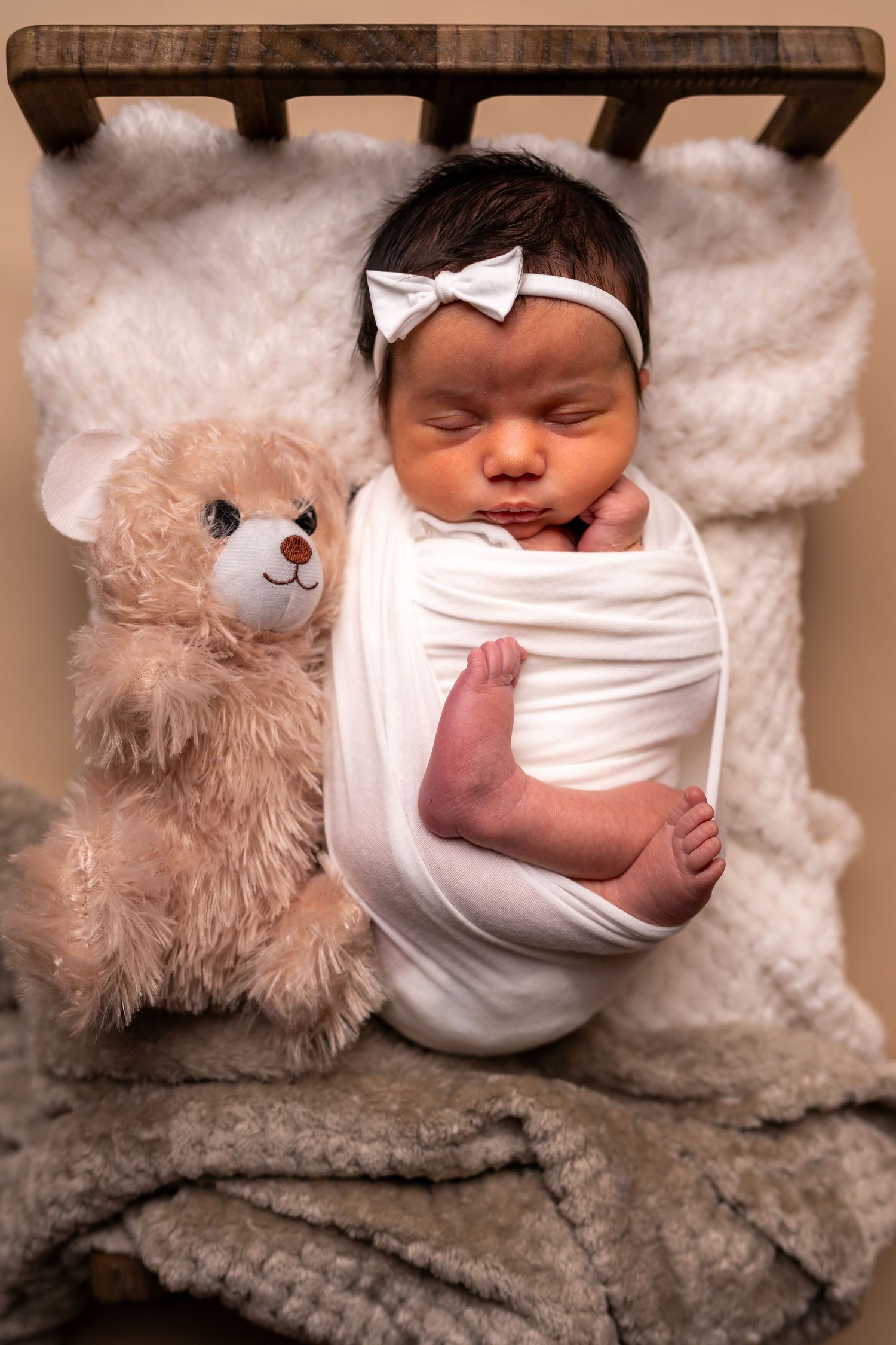 A sleeping newborn baby in white swaddle lies next to a plush teddy bear on a textured gray blanket.
