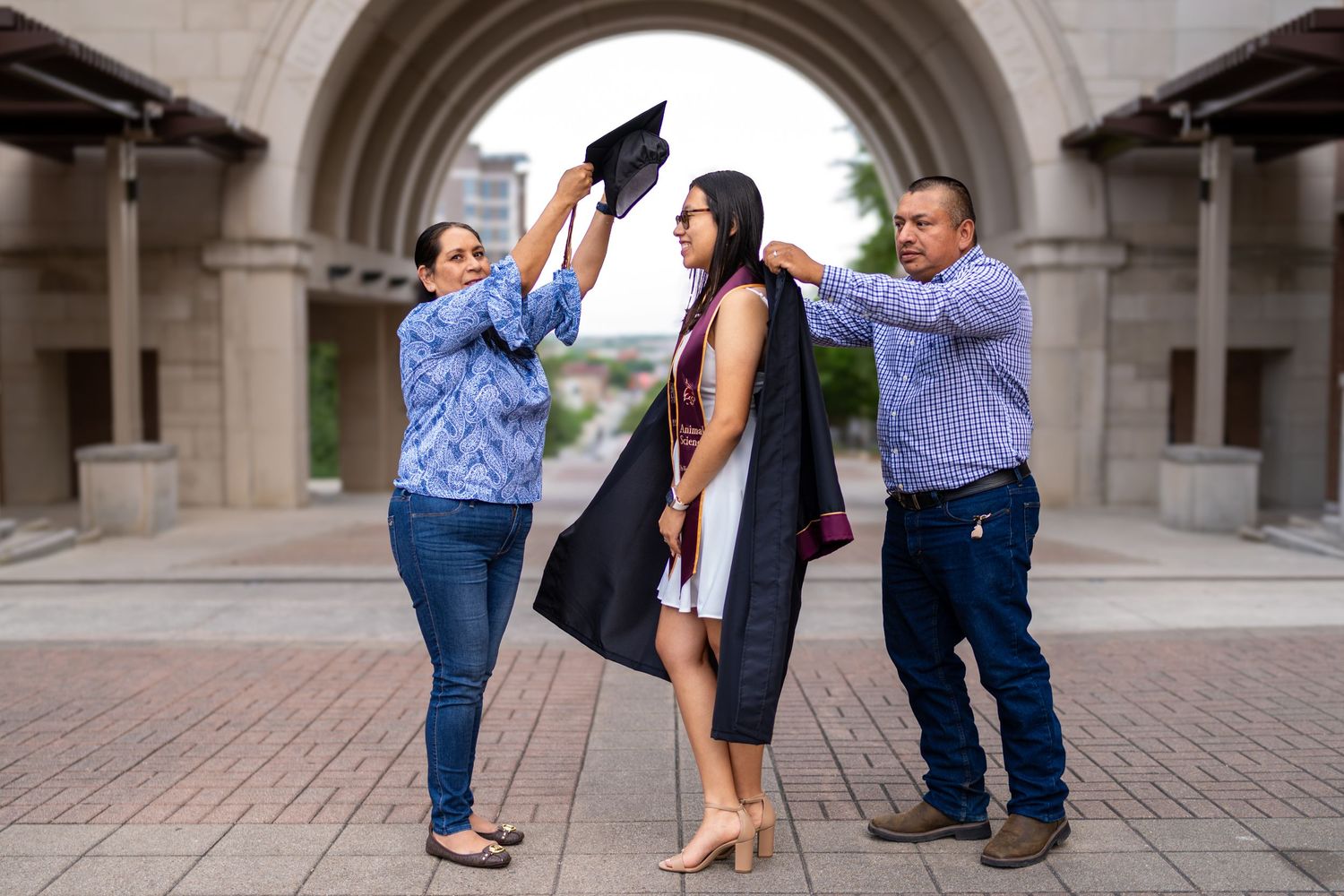 Parents proudly helping their daughter put on graduation cap and gown outdoors near stone archway.