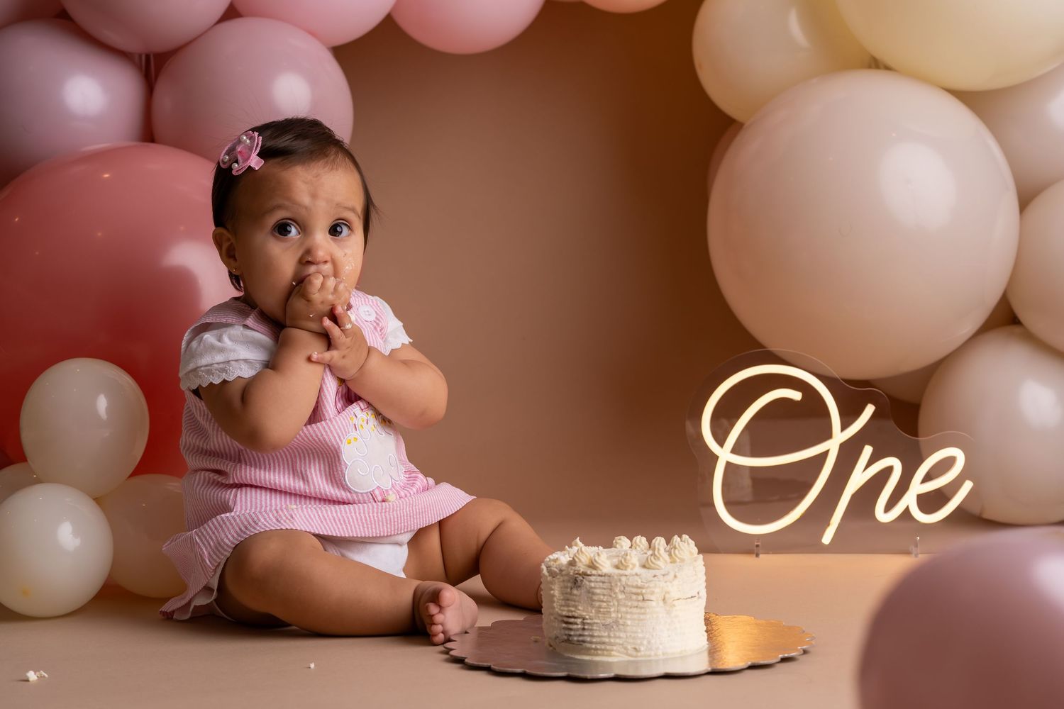 Baby in pink outfit sits among pink and white balloons next to a smash cake during first birthday photo session.
