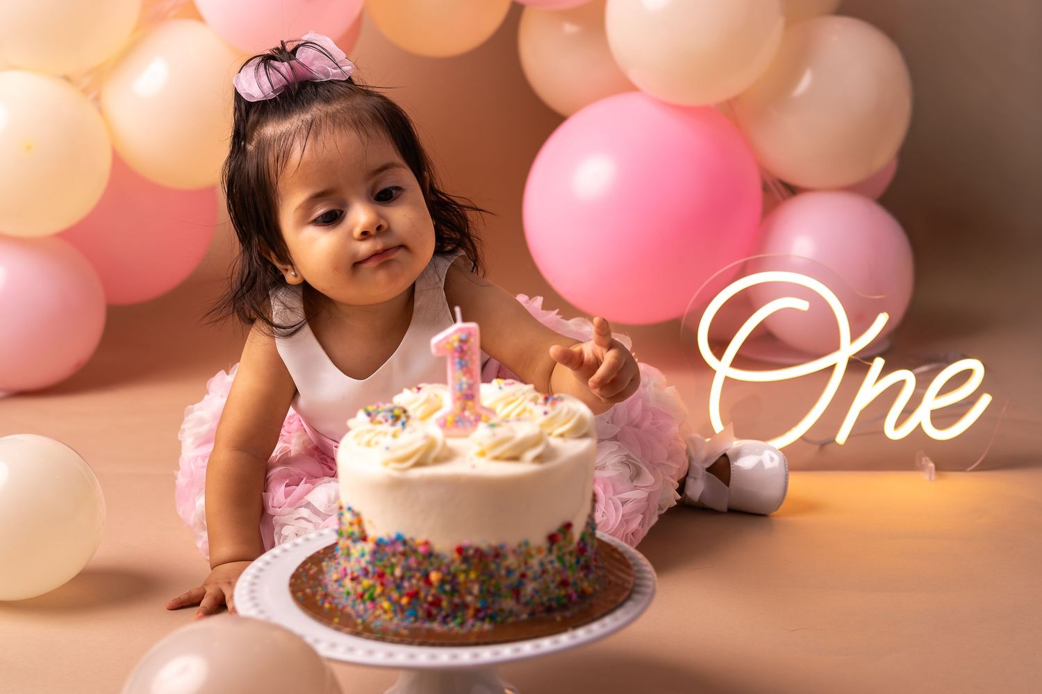 A pink and gold themed first birthday celebration featuring a chocolate cake with sprinkles and festive balloon decorations.