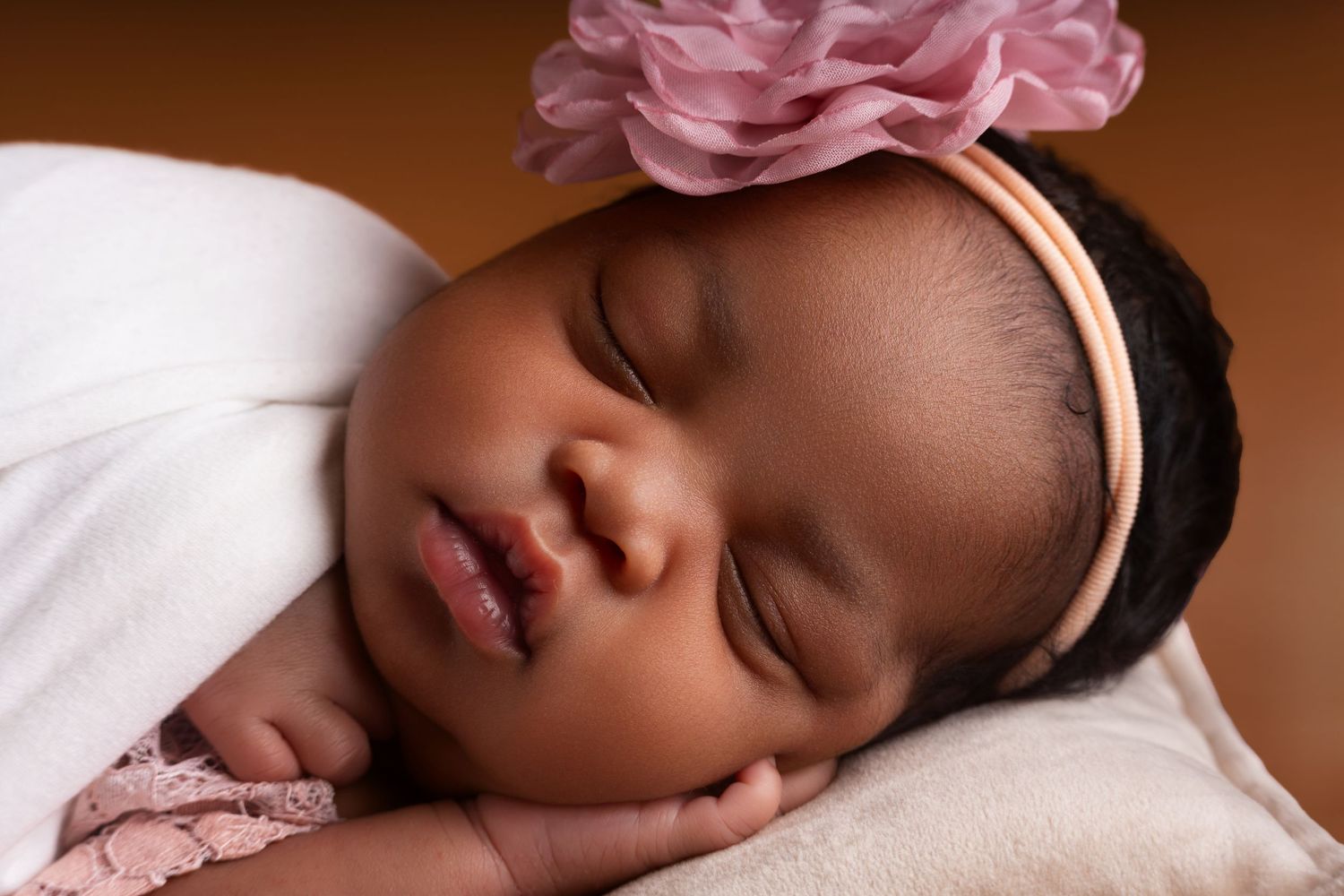 Sleeping newborn baby wearing a pink flower headband rests peacefully with head on tiny hand, wrapped in a soft white blanket.