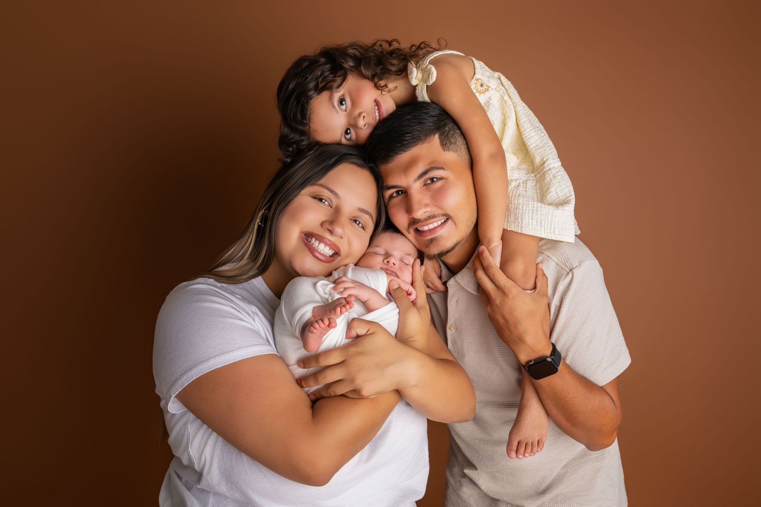 Smiling couple cuddling a newborn baby while a young girl sits on the father's shoulders in a warm family portrait against a brown backdrop.