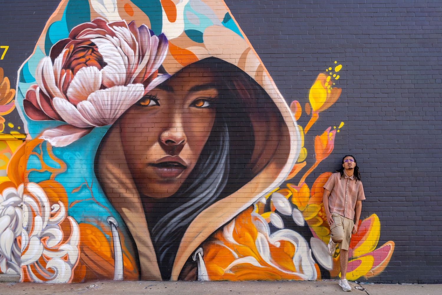 Woman stands beside vibrant street mural of hooded woman with floral crown on colorful orange and teal wall.