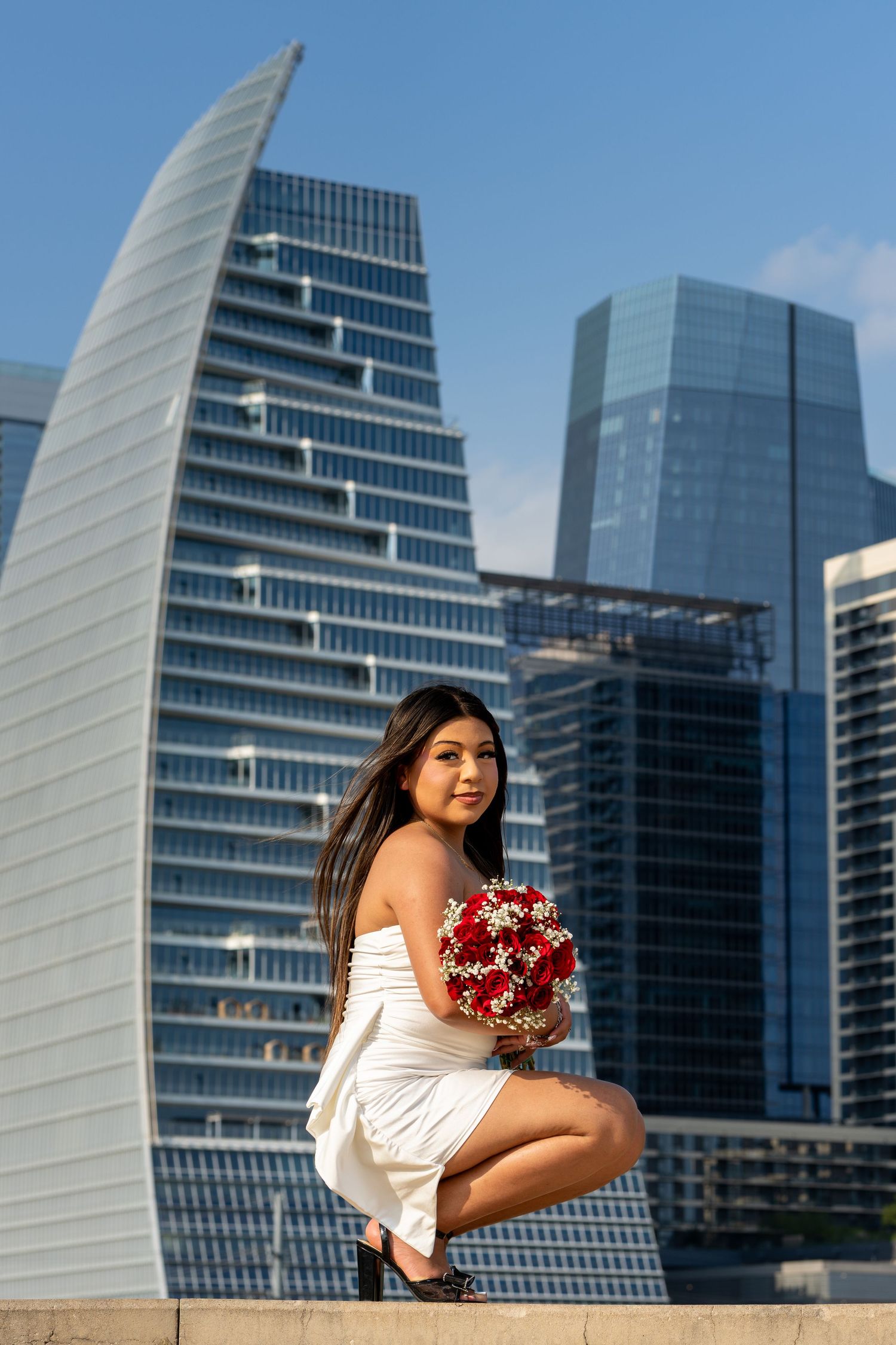 Young woman in white dress holding red bouquet poses before modern city skyscrapers on a sunny day.