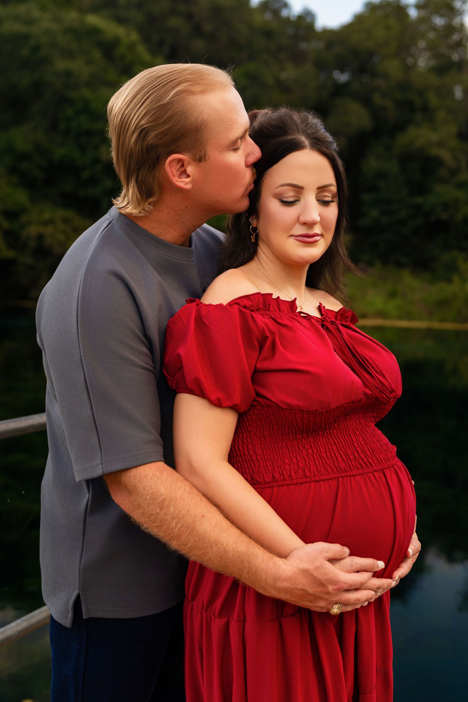 A man kisses a pregnant woman in a red dress while cradling her belly outdoors near a serene lake.
