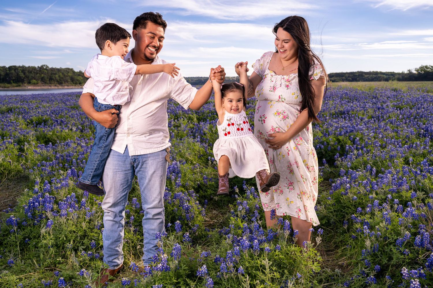 Pregnant woman and husband swing daughter in bluebonnet field while son watches, joyful family maternity photo in Texas.