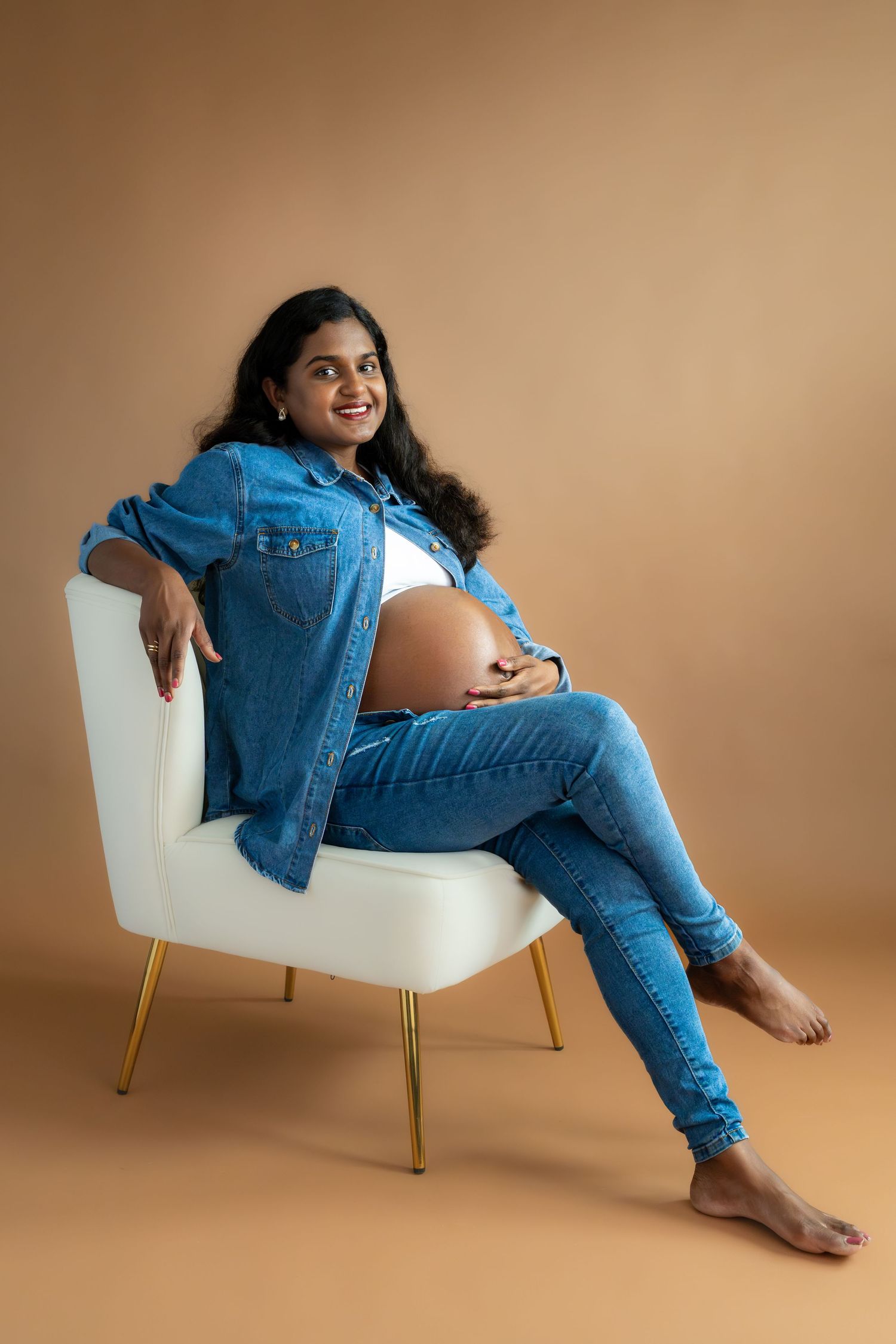 Smiling pregnant woman in denim jacket and jeans sitting in white chair, cradling baby bump against beige background.