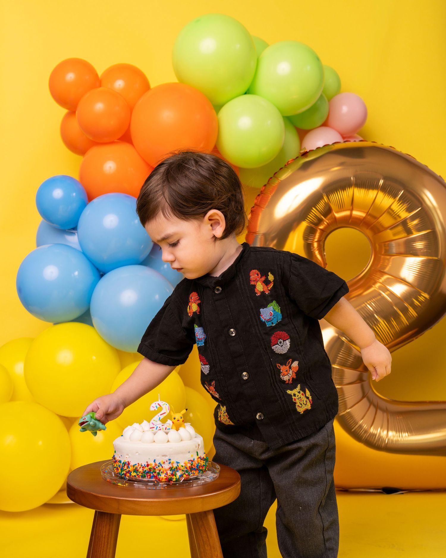 Birthday celebration with colorful balloons, gold number 2, and a cake on a wooden table against yellow backdrop.