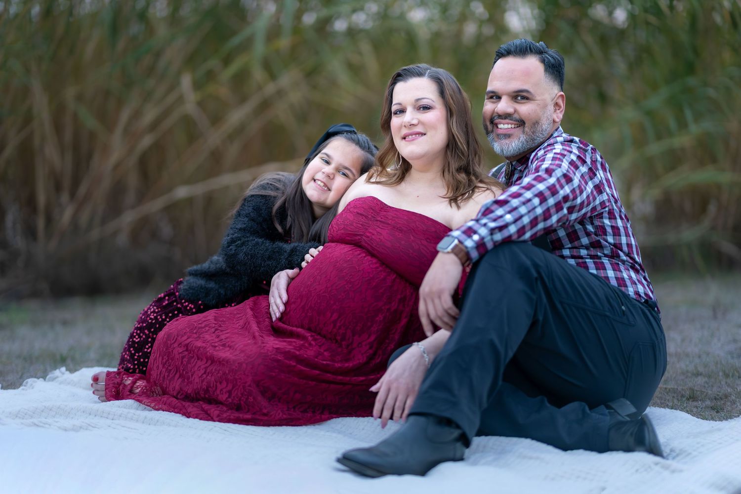A family maternity portrait on a sandy beach with everyone wearing coordinating burgundy and plaid outfits.