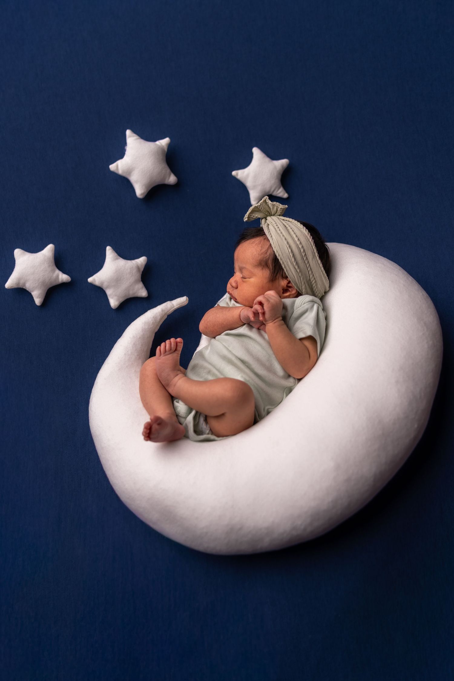 Sleeping newborn rests peacefully on white crescent moon prop with decorative stars against dark blue backdrop.
