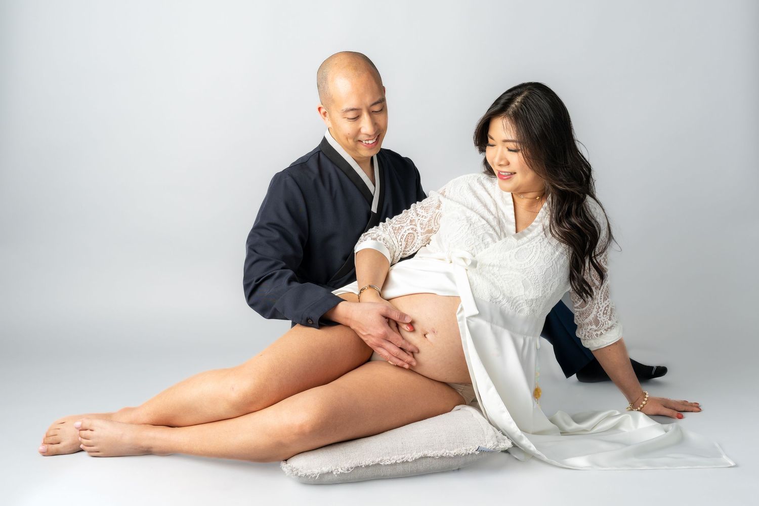 Couple in maternity photoshoot, man tenderly touching pregnant woman's belly, seated on white floor.