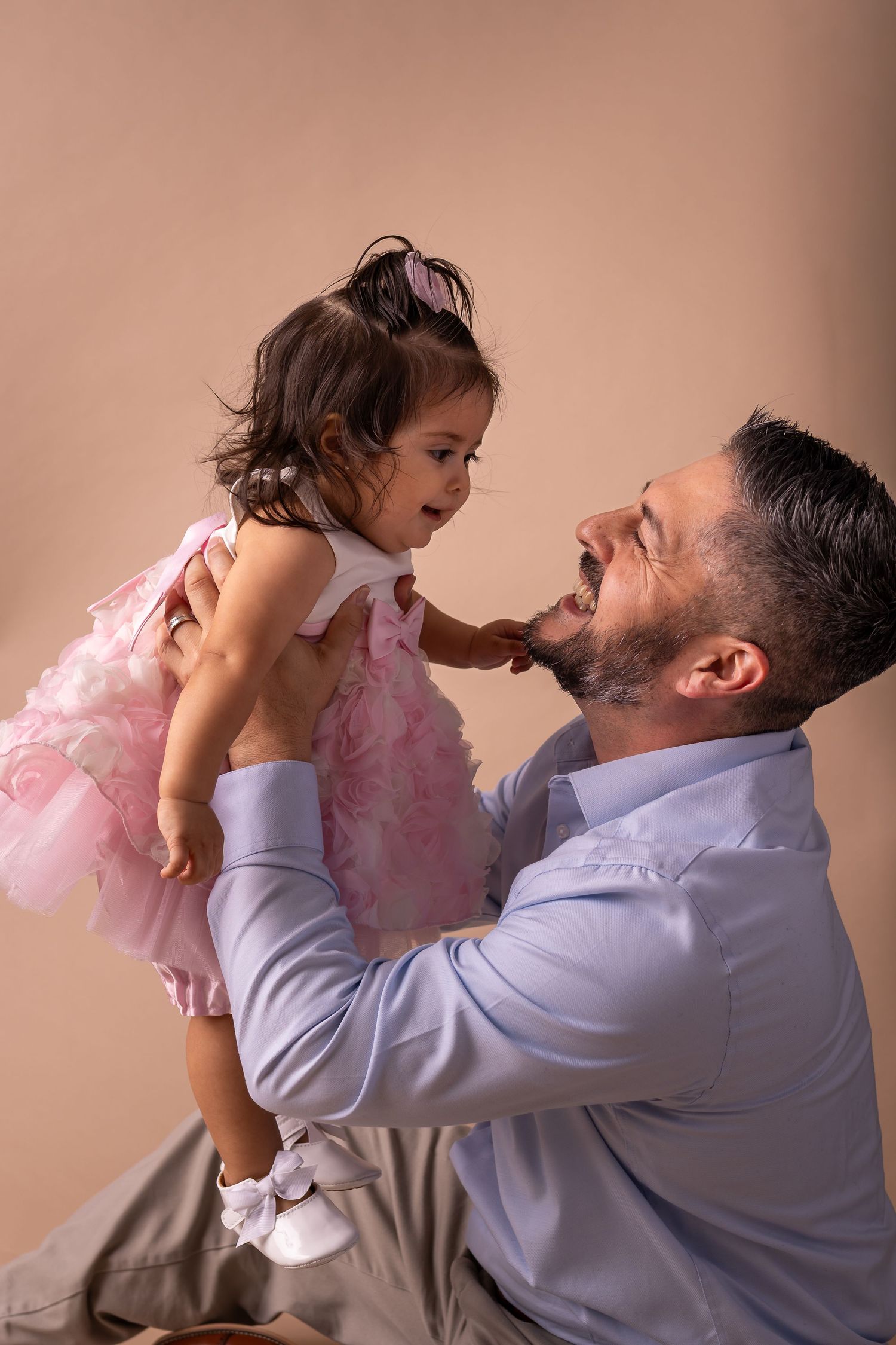 A parent in a blue shirt lifts and plays with a small child wearing a pink tutu dress against a beige background.