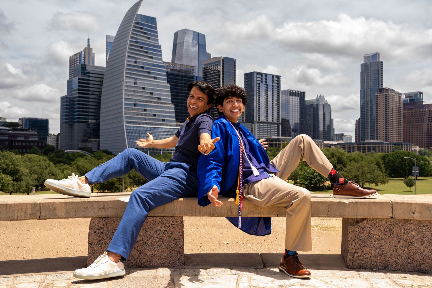 Two graduates in blue robes pose back-to-back on a ledge with the Austin, Texas skyline behind them.