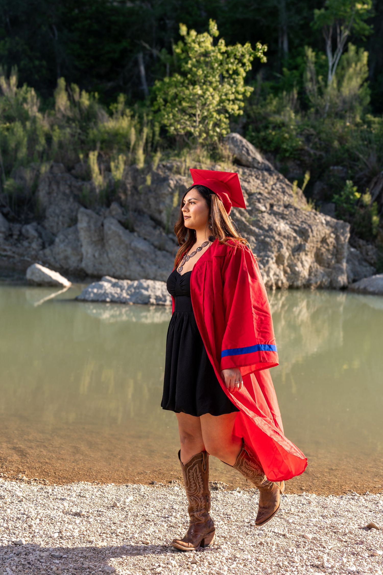 Graduate in red cap and gown walks along rocky lakeside wearing cowboy boots and black dress.