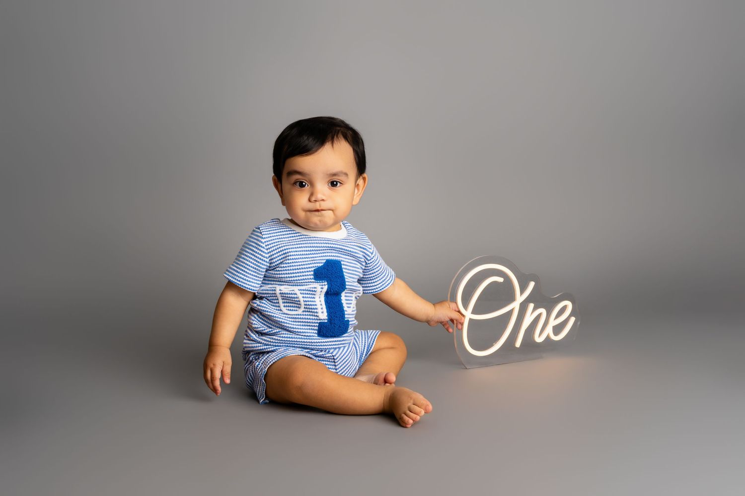 Baby boy in blue outfit sits beside a white 'One' sign on gray backdrop, celebrating his first birthday.