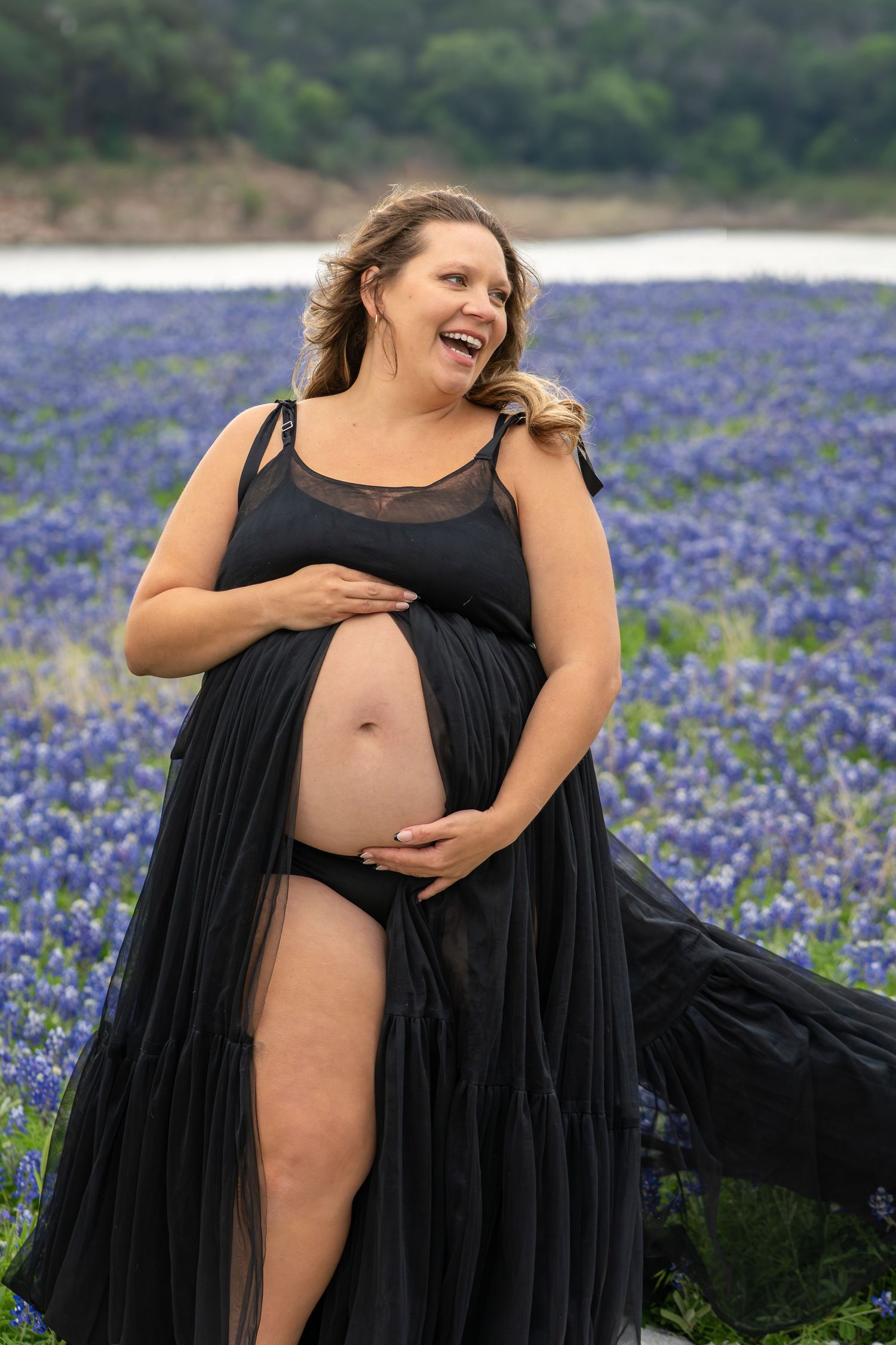 Pregnant woman in black maternity gown laughing in a Texas bluebonnet field, cradling her baby bump.
