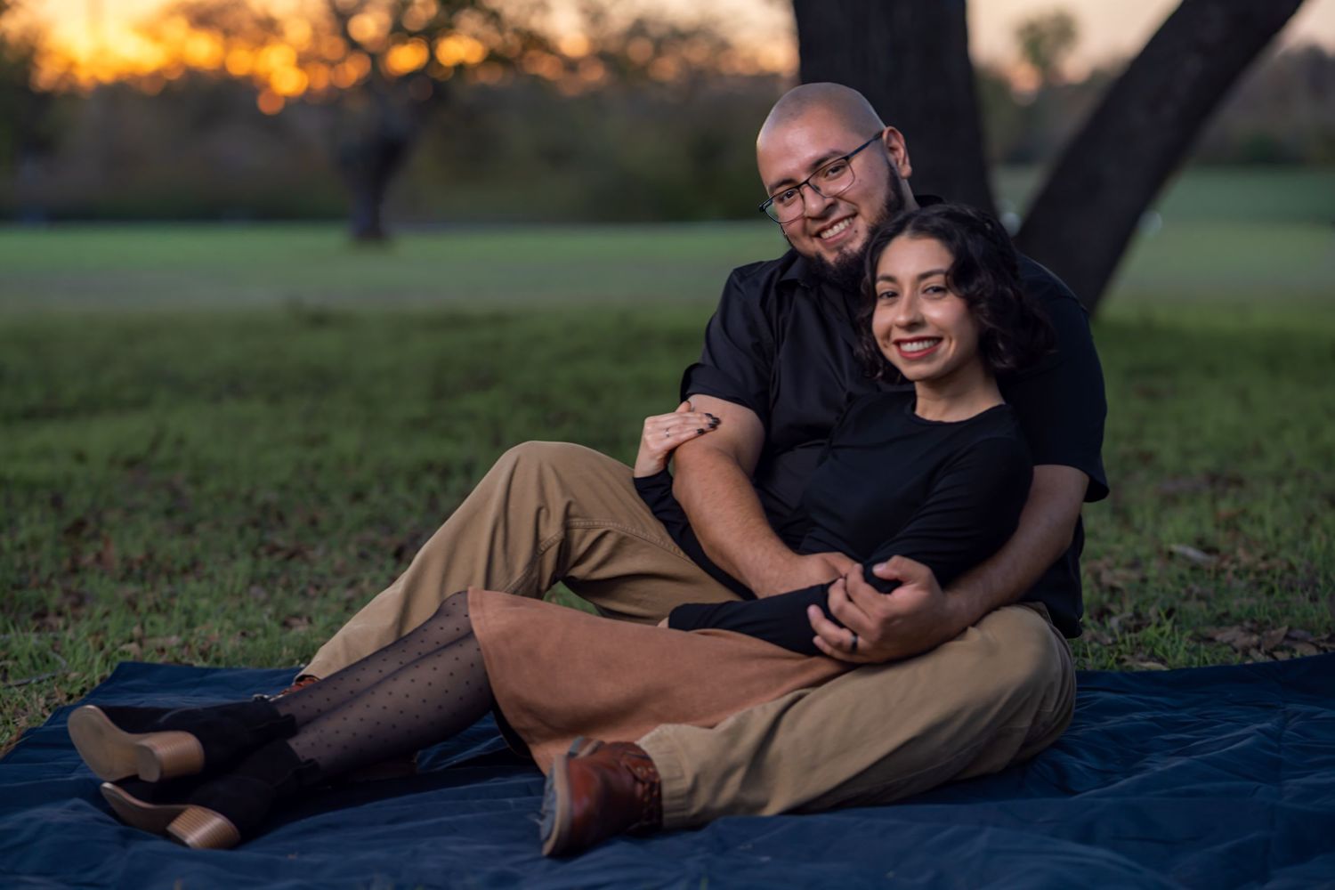 A couple shares an intimate moment while lying together on a blanket in a grassy outdoor setting at sunset.
