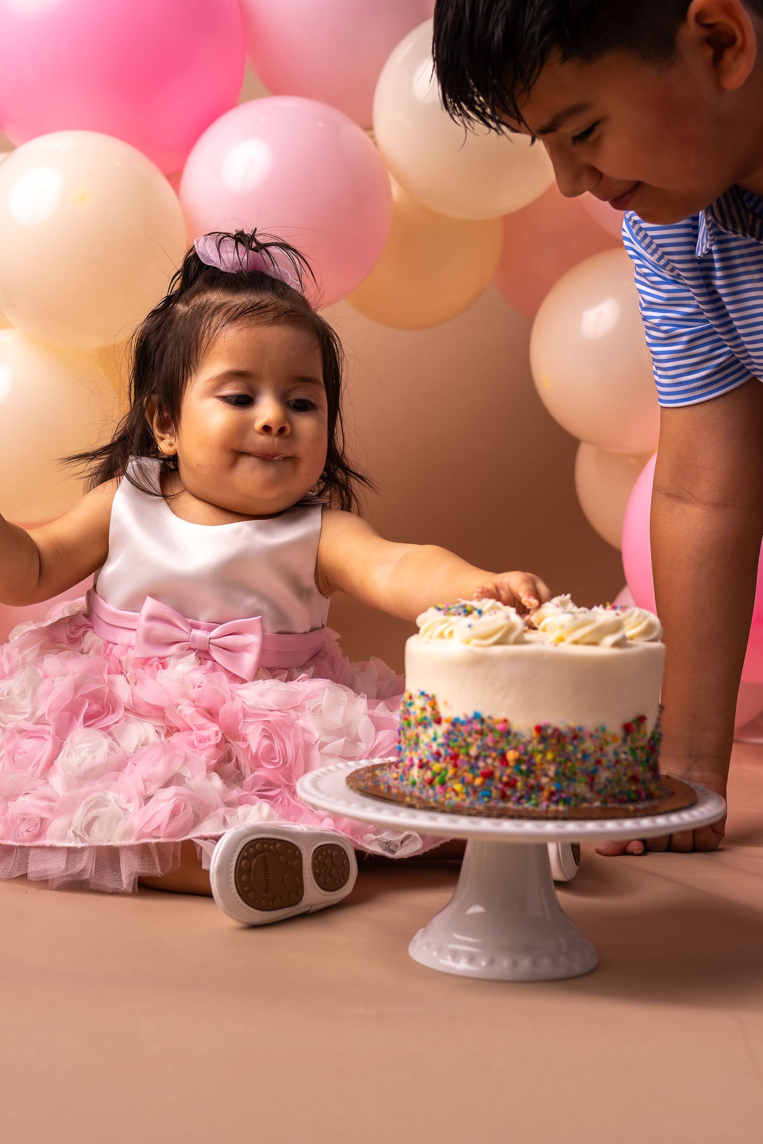 A cake smash photo session showing a baby in a pink tutu dress reaching for a white frosted birthday cake with sprinkles.