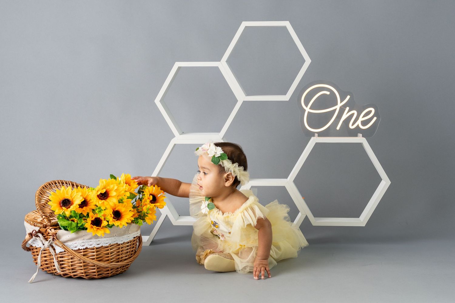 Baby girl in yellow tutu touches sunflowers in basket, sitting before honeycomb decor with One sign, first birthday photo.