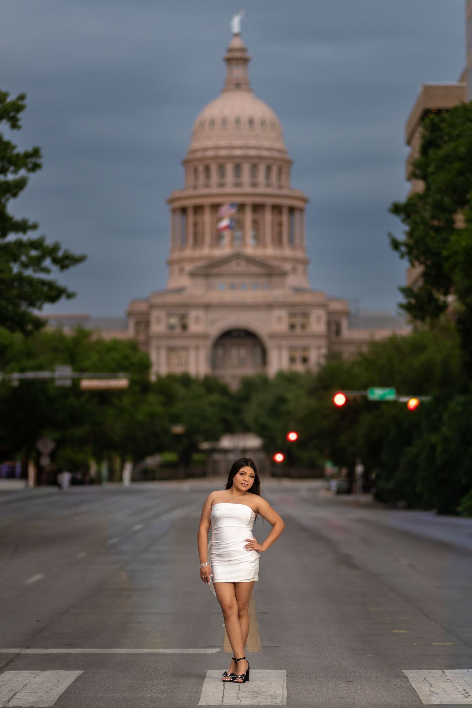 Woman in white dress poses on street in front of Texas State Capitol building at dusk.