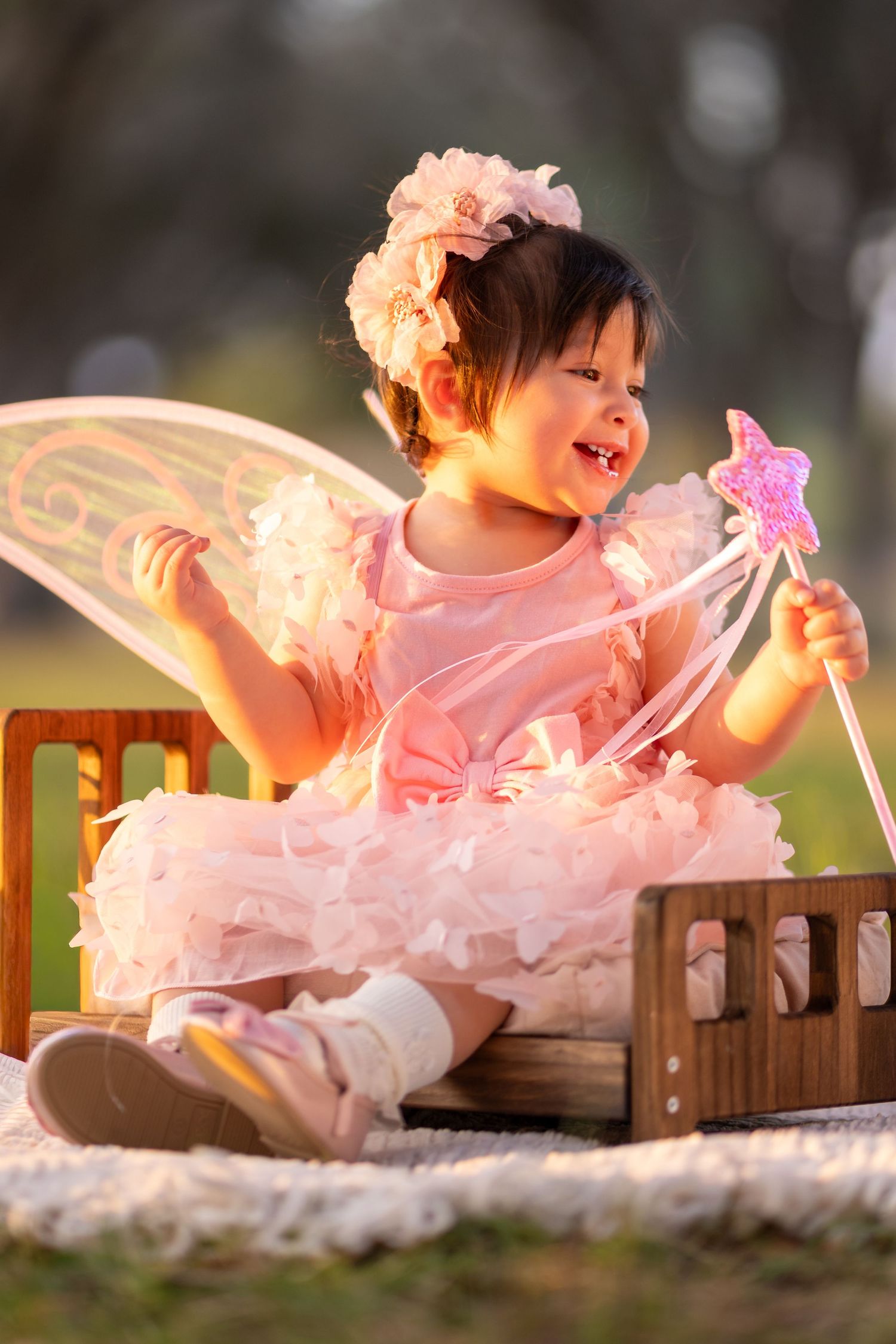 Happy toddler in pink tutu fairy dress holds magic wand while sitting outdoors in golden light.