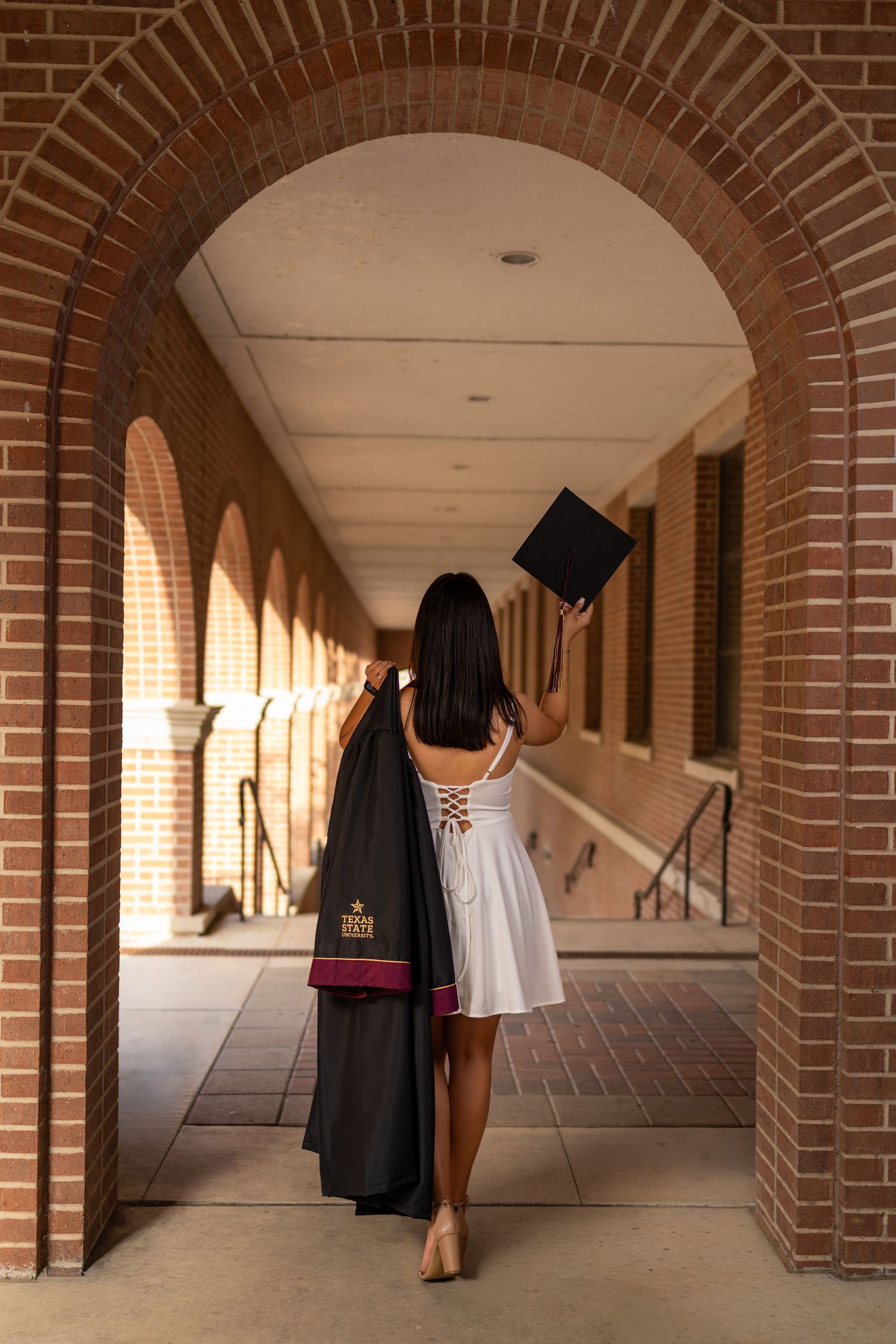 Graduate in white dress holds cap and gown under brick archway colonnade, celebrating academic achievement.