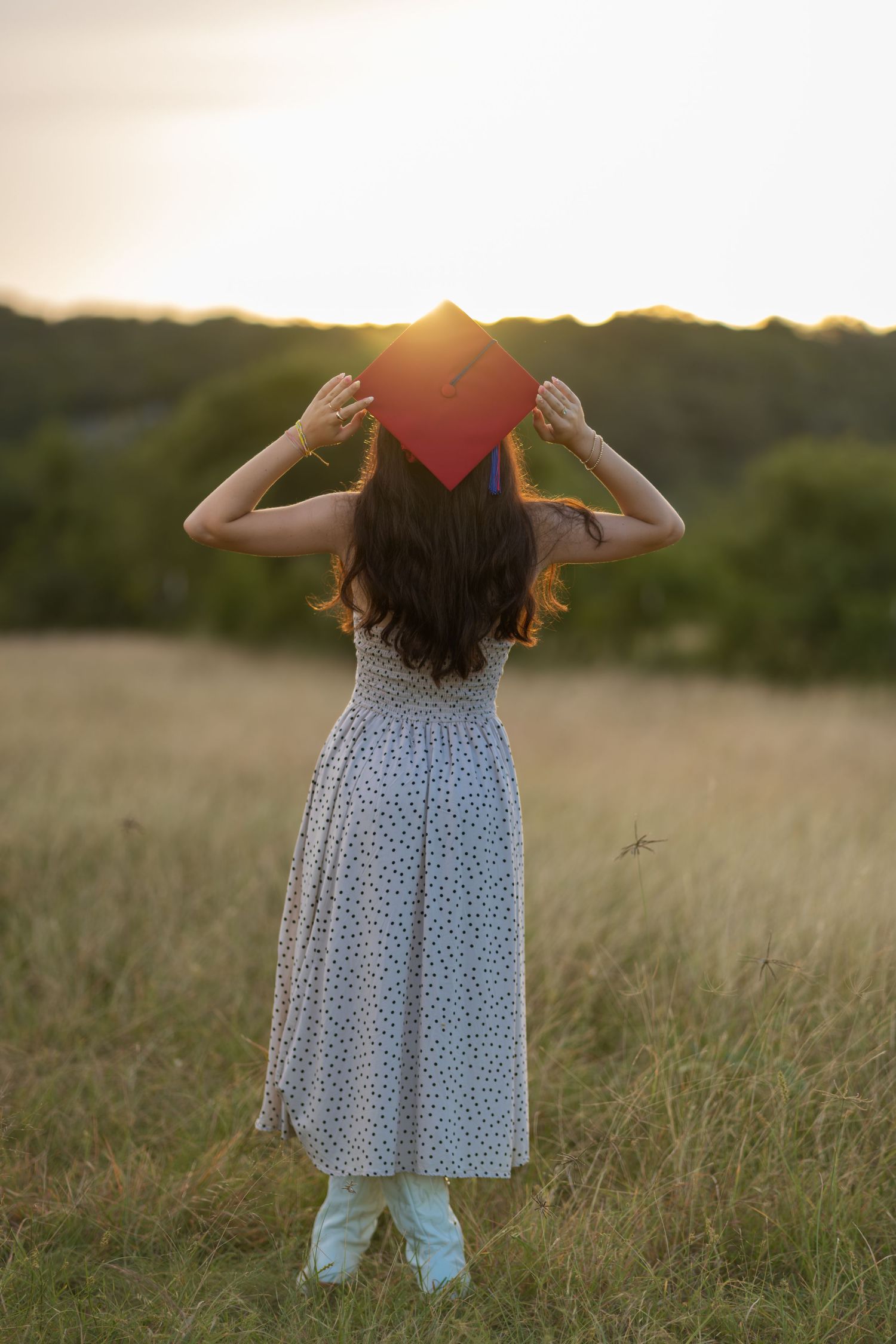 Girl in floral dress holding red graduation cap in golden field at sunset, viewed from behind.