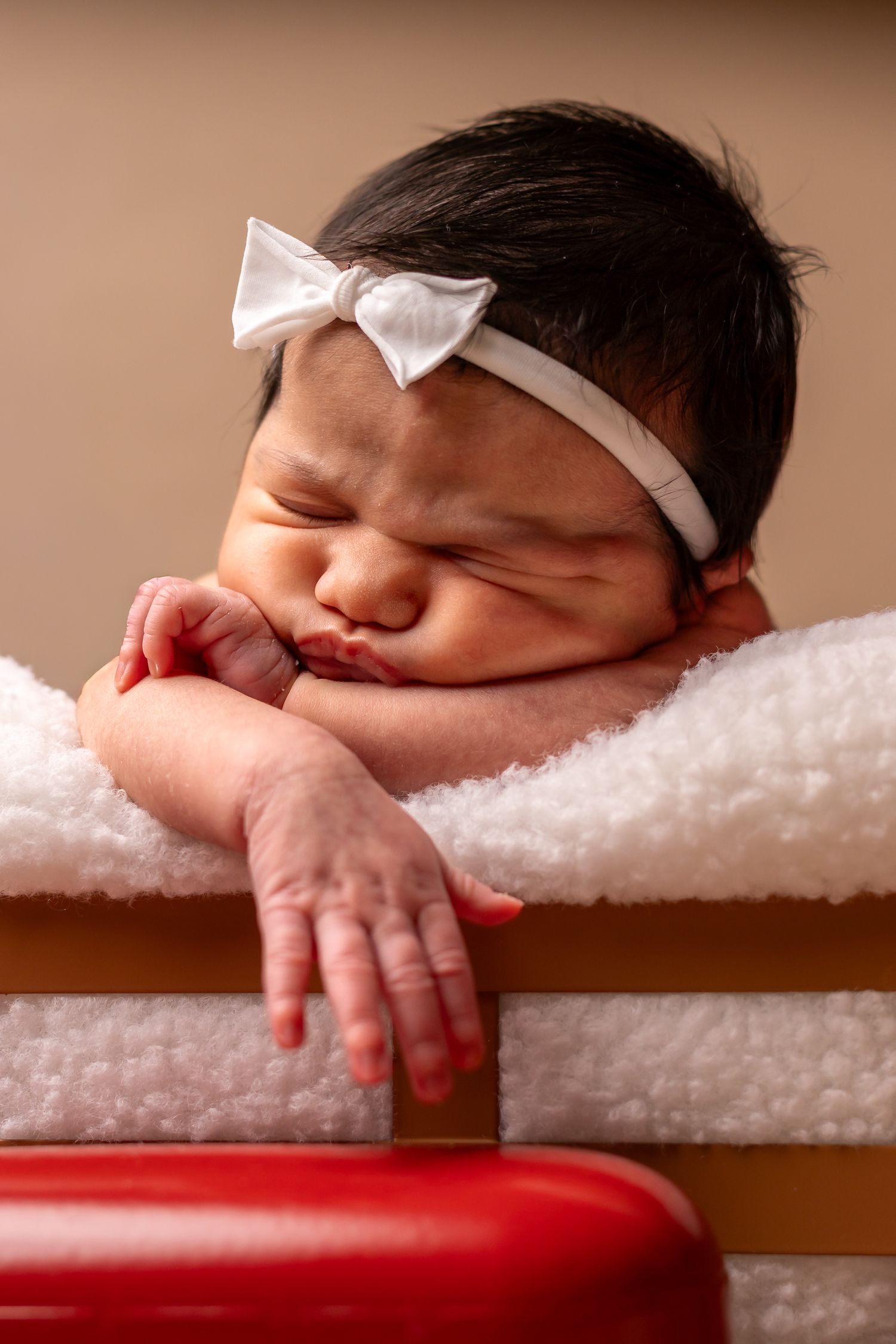 Sleeping newborn in white bow headband rests peacefully on soft white towels next to red surface.