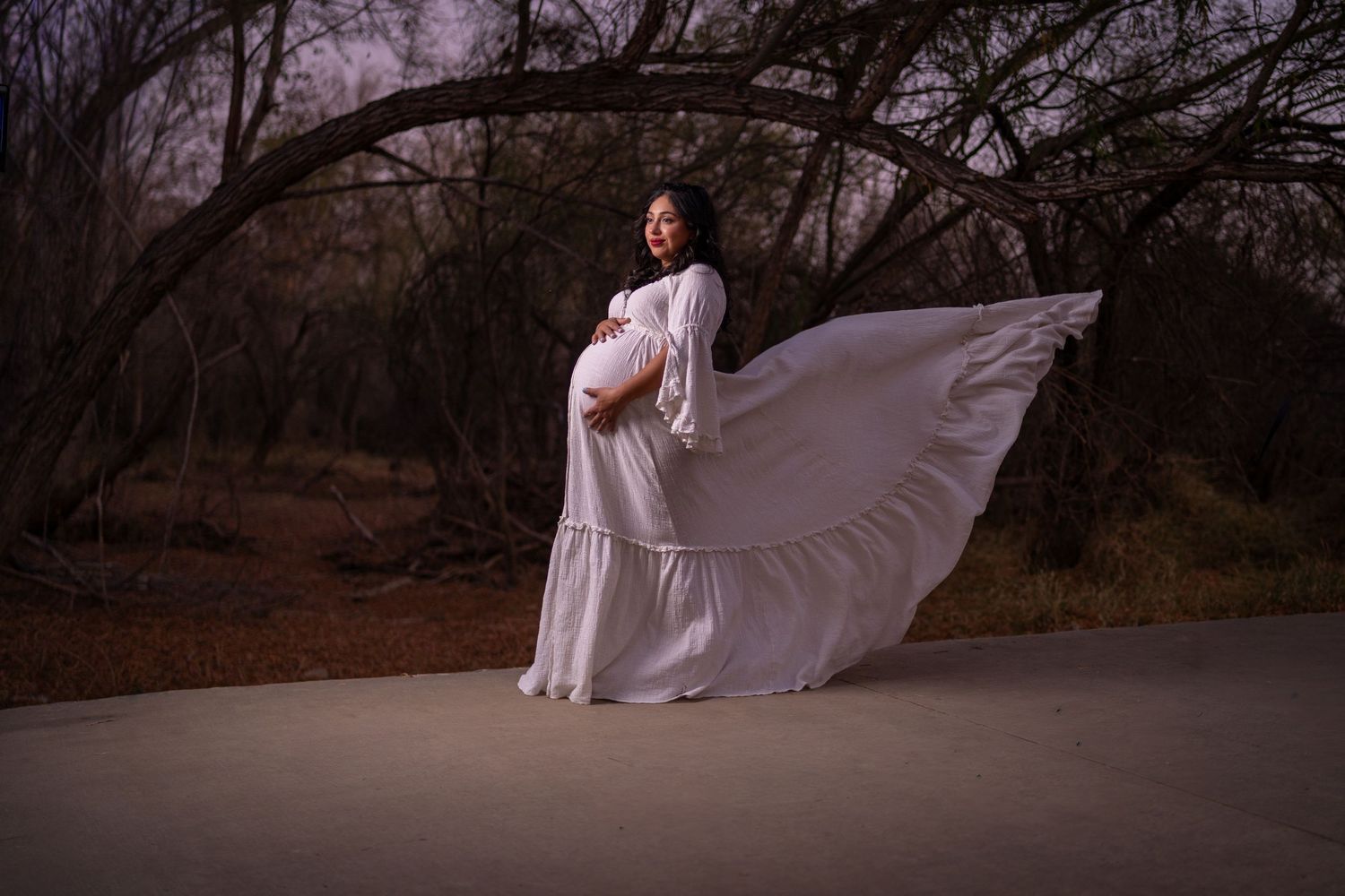 A flowing white dress billows dramatically on a path surrounded by bare winter trees at dusk.
