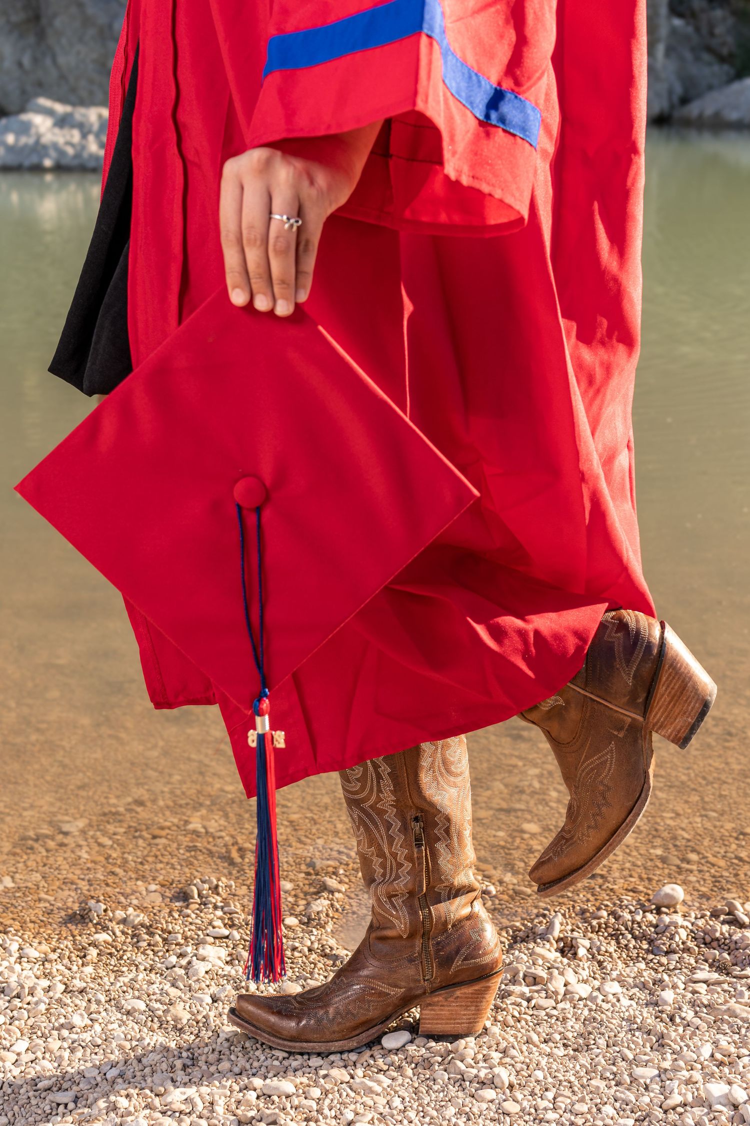 Close-up of a graduate in red gown and cowboy boots holding a red mortarboard cap with tassel.